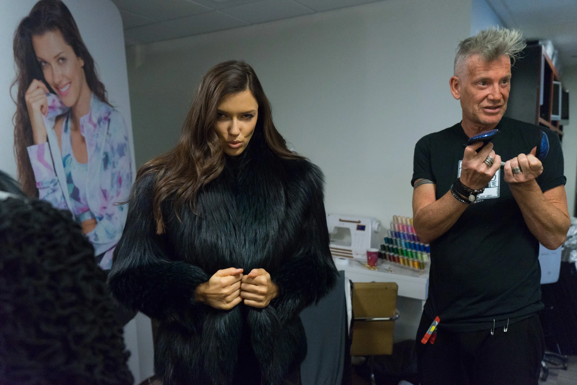 A model in a black textured coat standing beside a person gesturing with both hands.