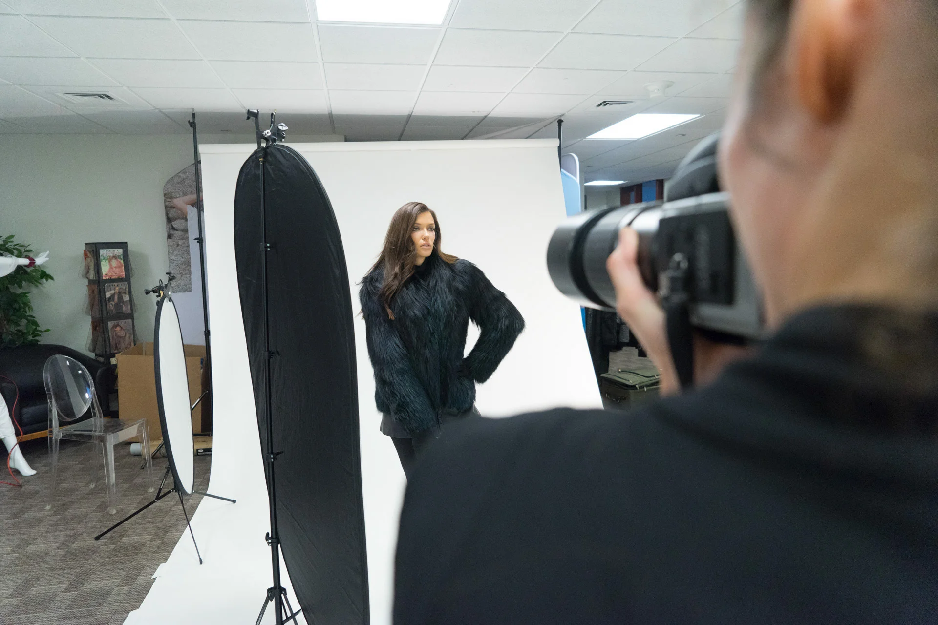 A photographer framing a model in a black coat on a white studio backdrop.