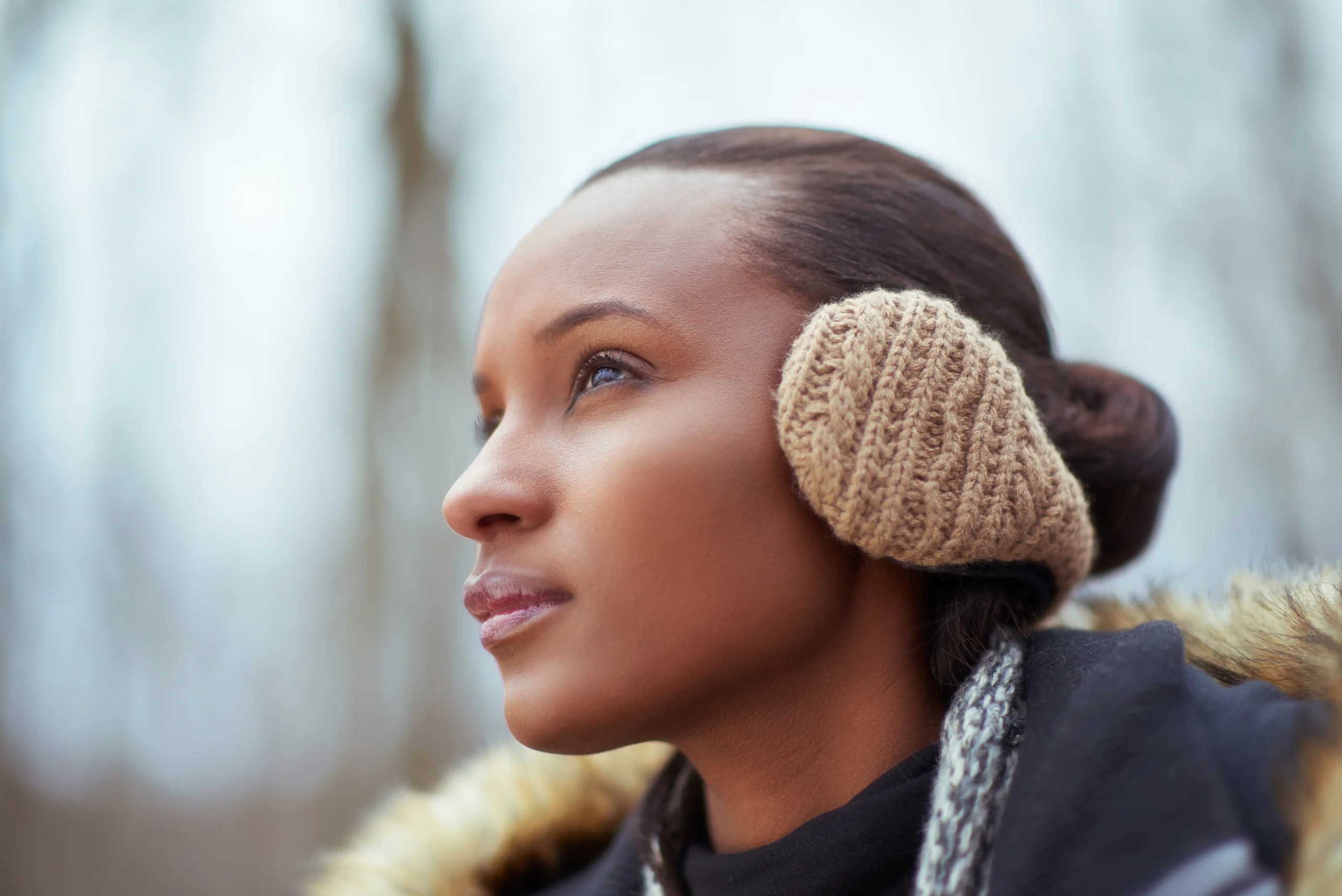 A close outdoor portrait of a model wearing beige knitted earmuffs.