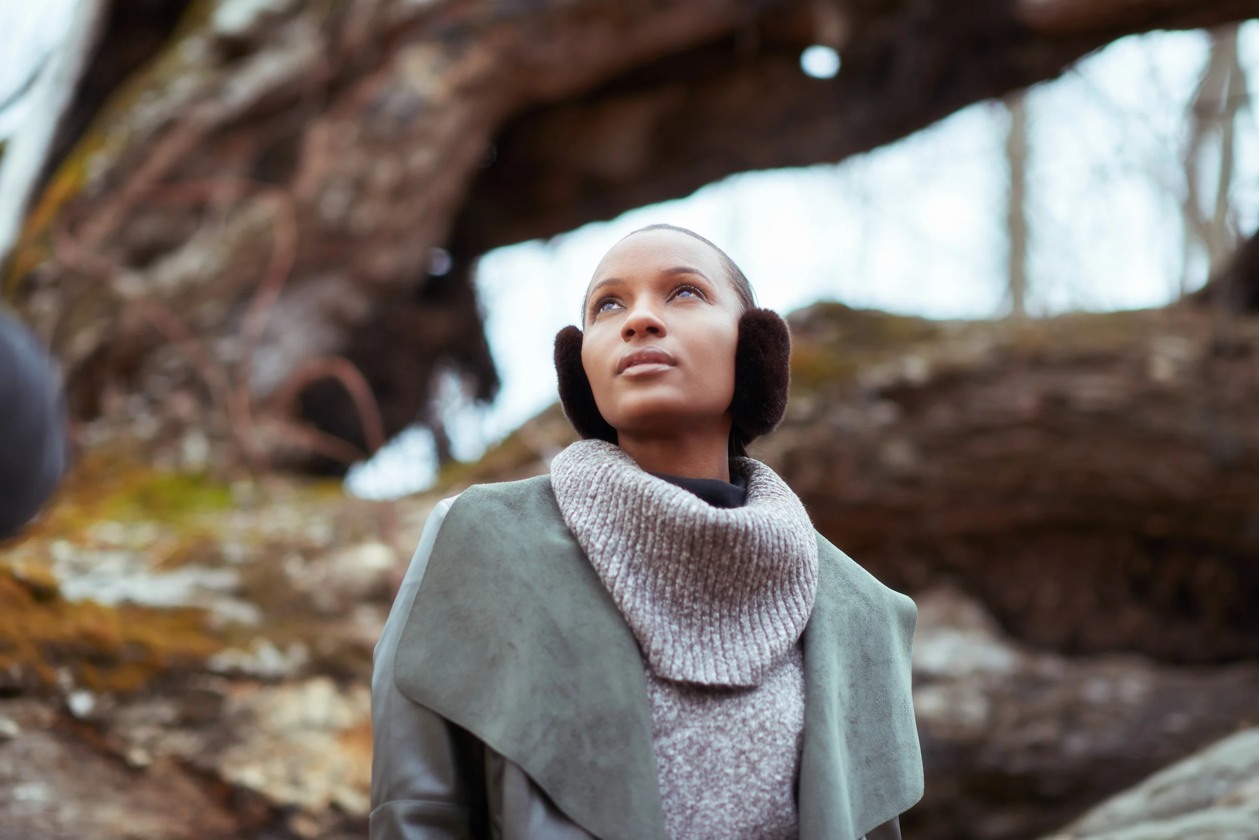 A model wearing dark earmuffs and a gray cowl-neck sweater beneath a fallen tree.