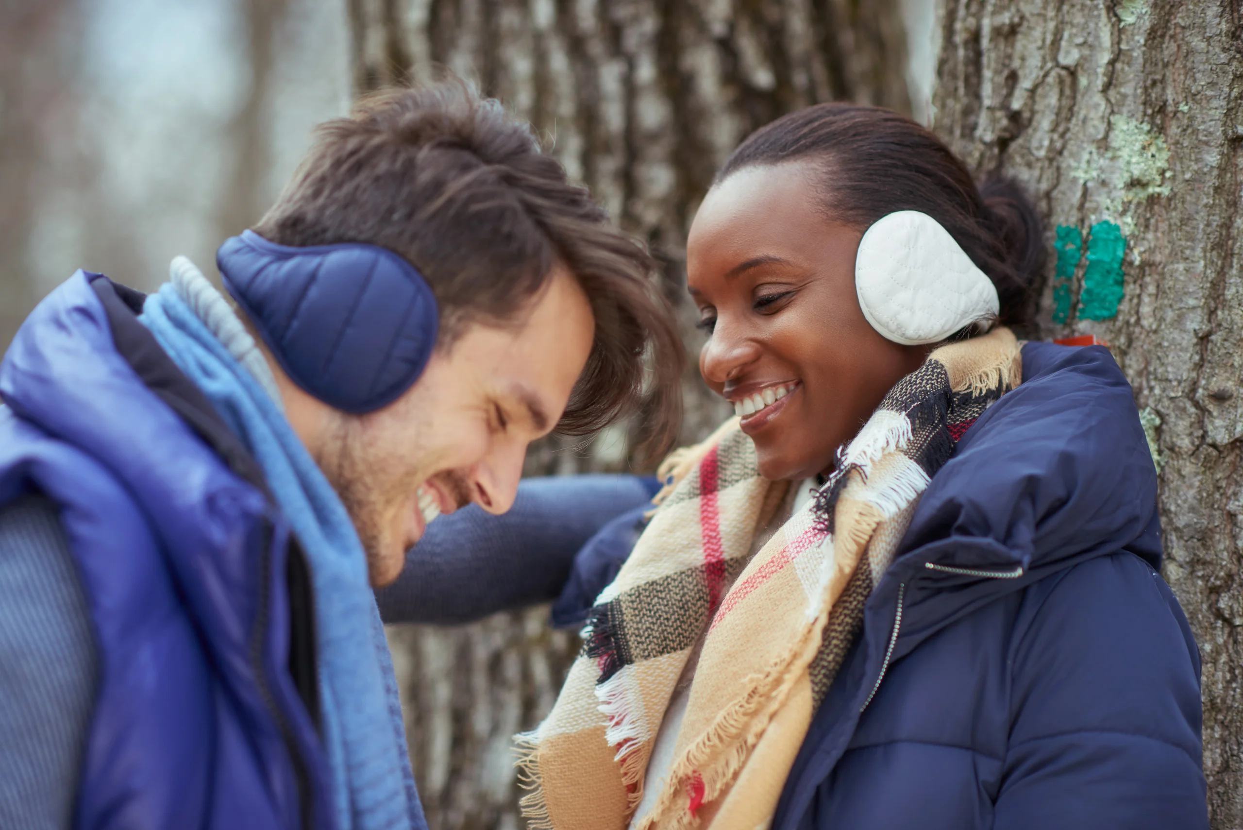 Two smiling models in winter coats and earmuffs standing beside a tree.