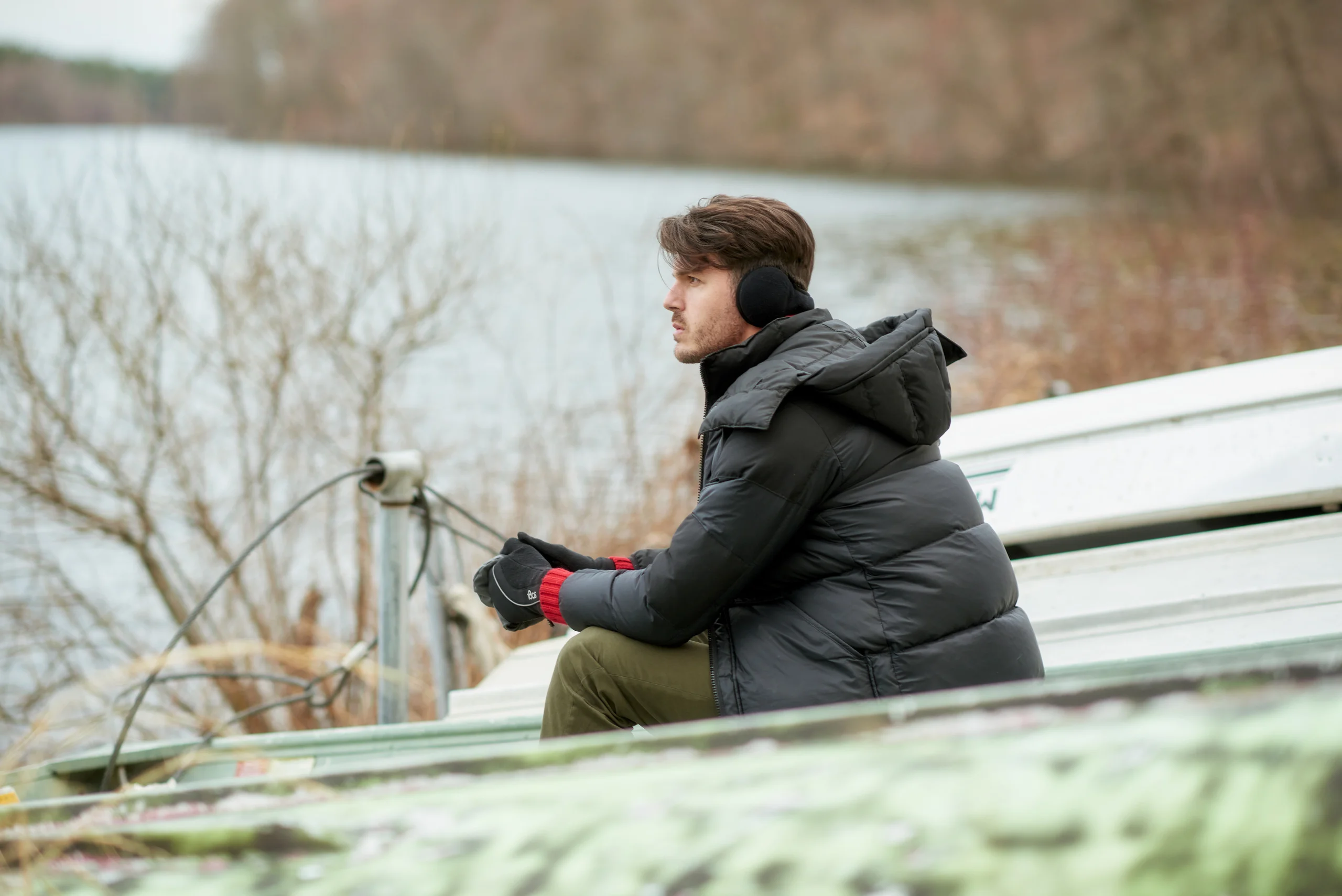 A model in a dark padded coat sitting on a bench beside water.