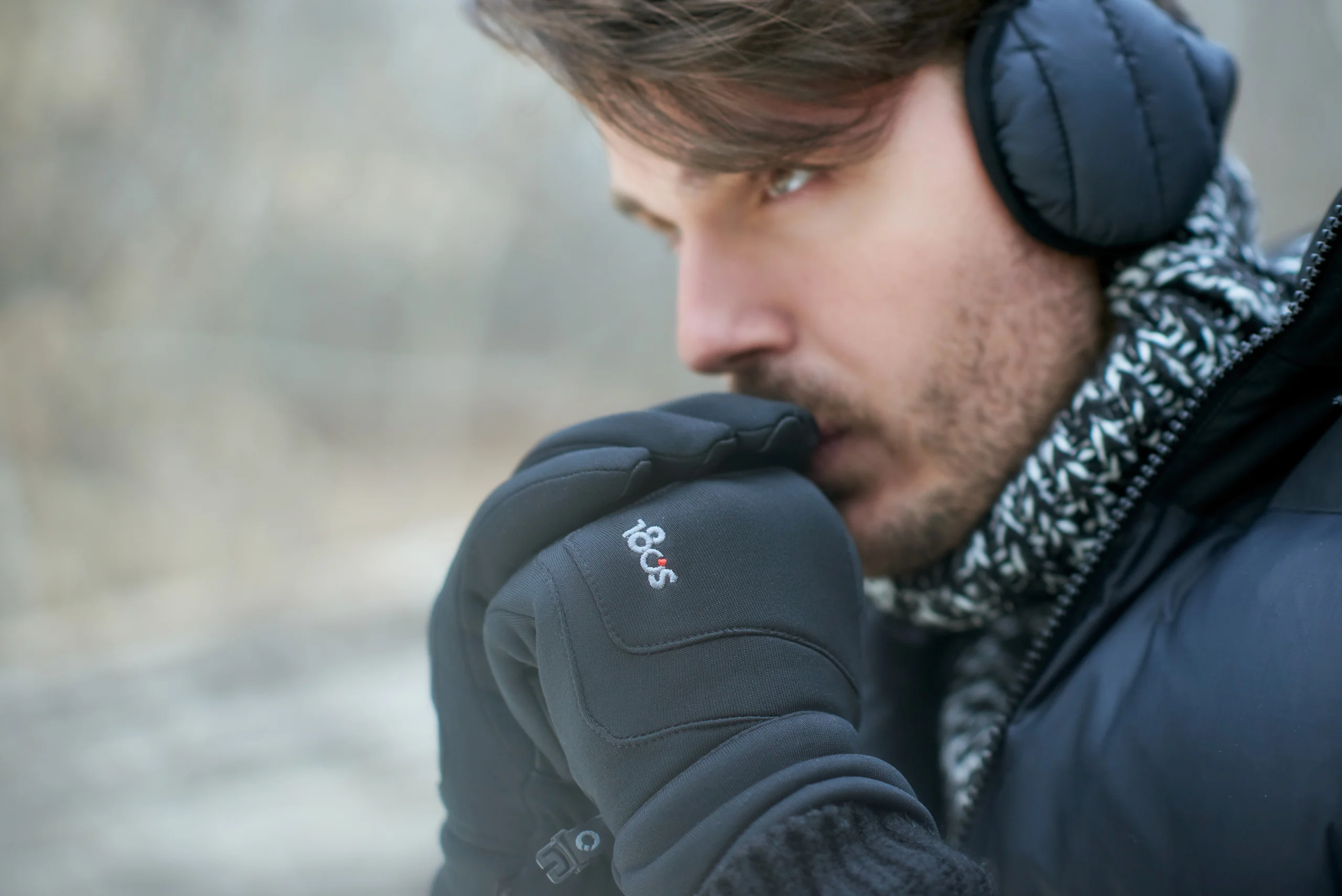 A close portrait of a model holding black-gloved hands near the face beneath dark earmuffs.