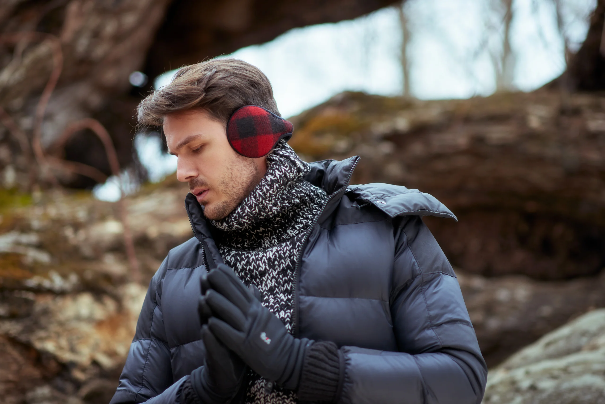 A model in red-and-black earmuffs holding gloved hands together beside a fallen tree.