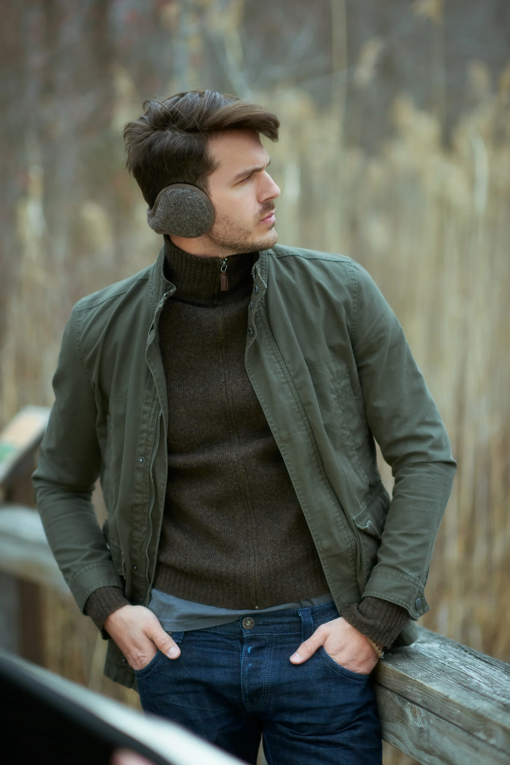 A model in a green jacket and gray earmuffs standing beside a wooden railing.