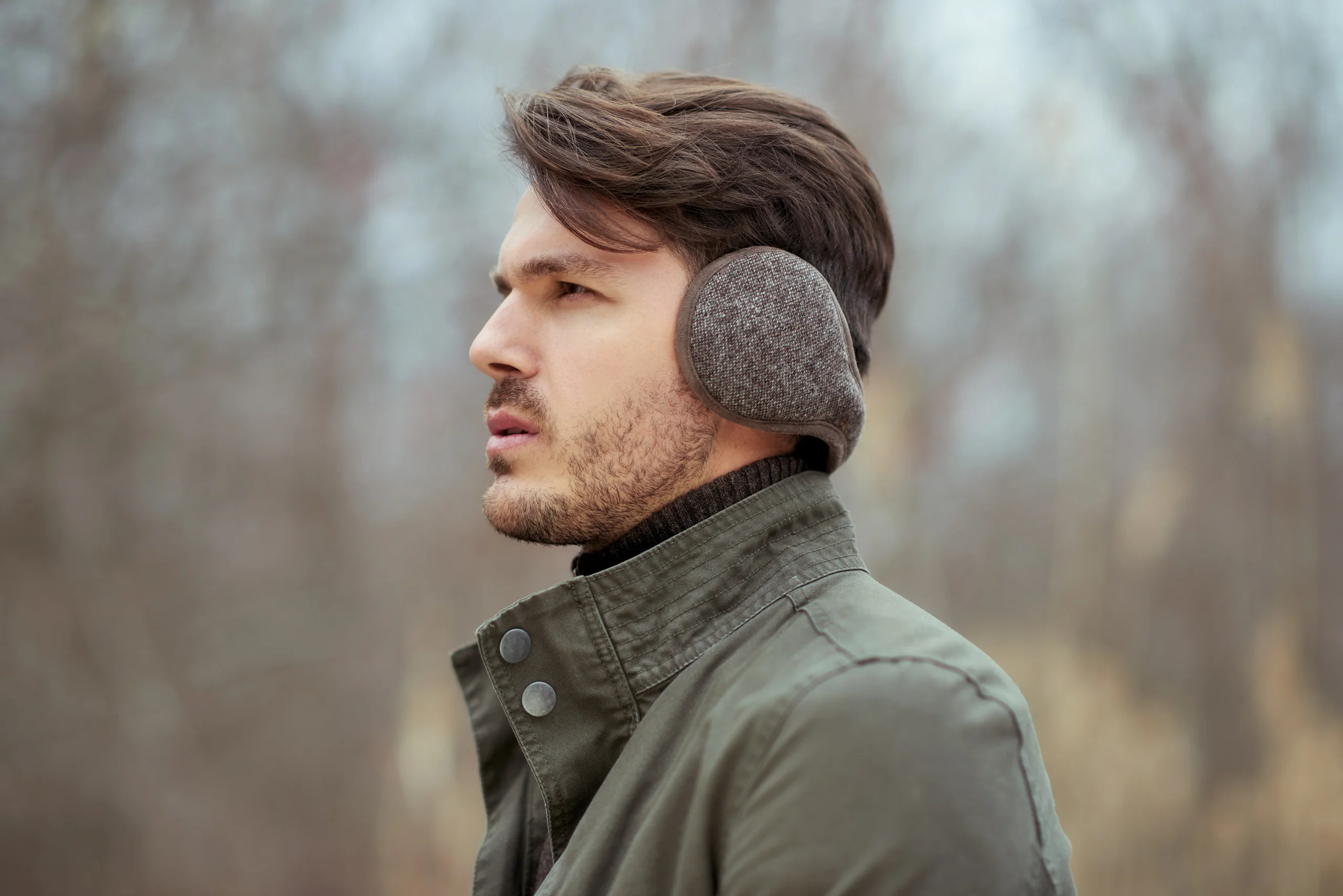 A side portrait of a model wearing gray knitted earmuffs and a green jacket.