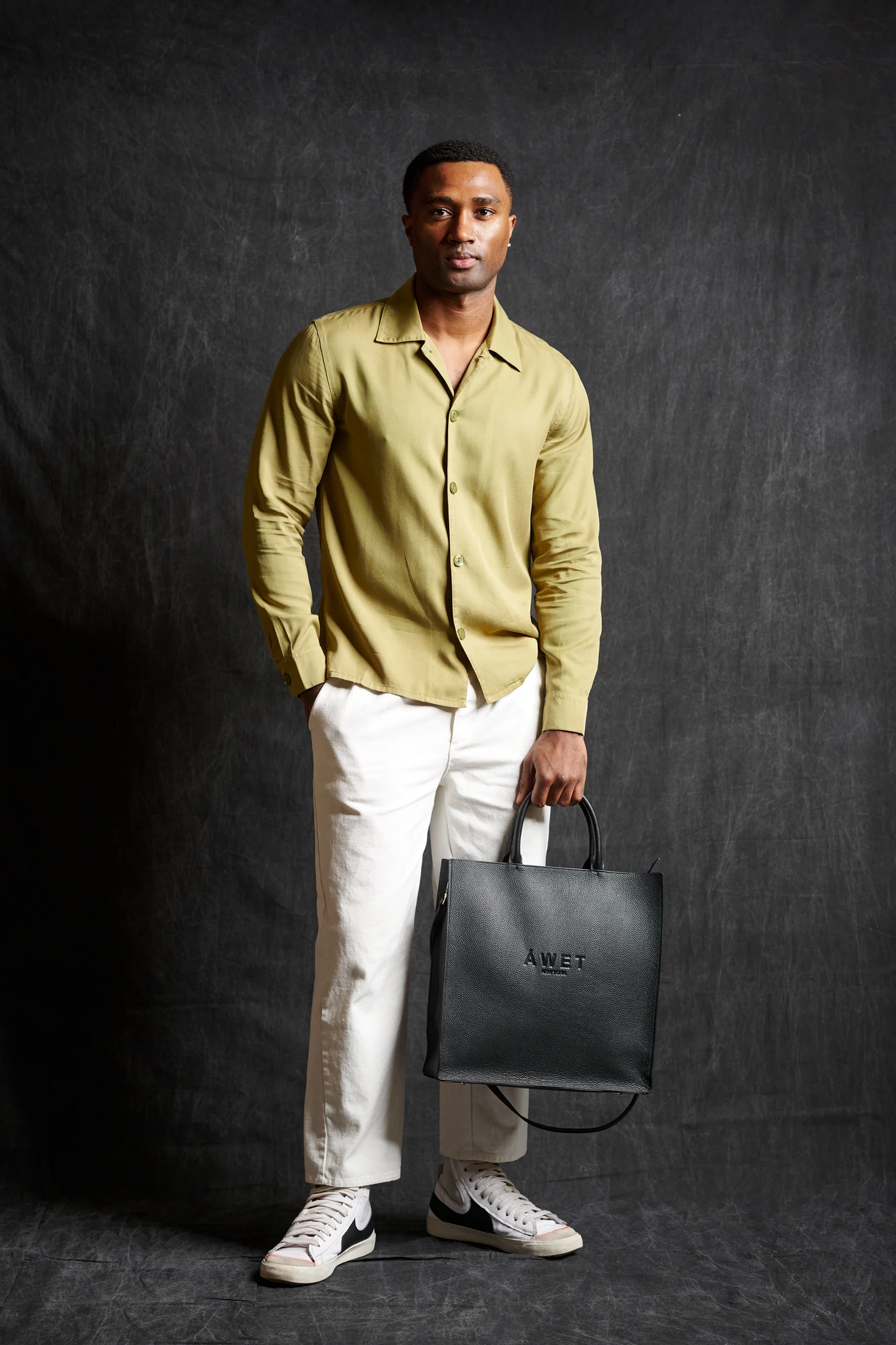 A model wearing an olive shirt and white trousers, holding a black bag.