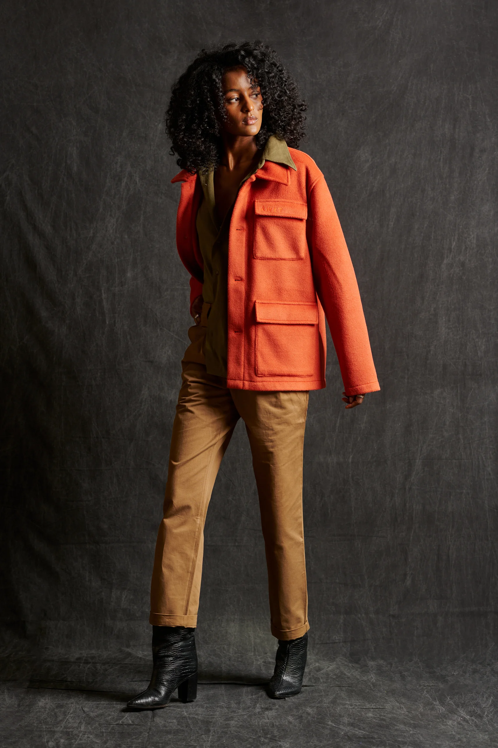 A model in an orange jacket, tan trousers and black boots against a charcoal backdrop.