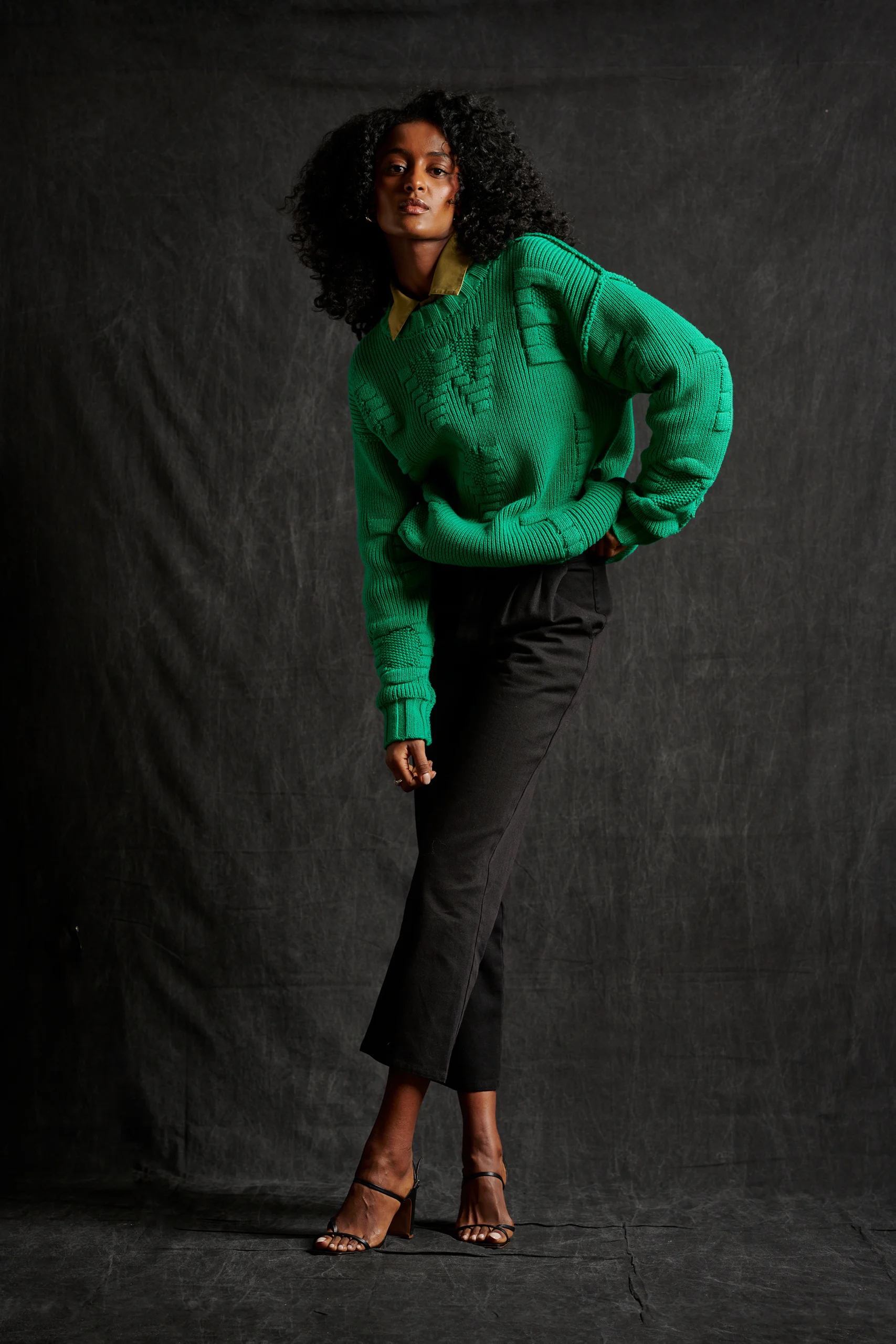 A model in a green sweater leaning forward with one hand on the hip.