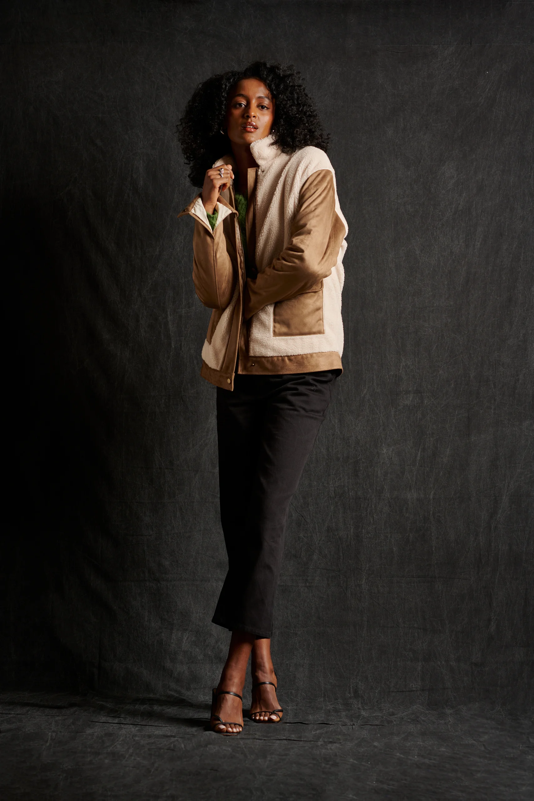 A model in a tan-and-cream cardigan and cropped black trousers, with one hand near the face.