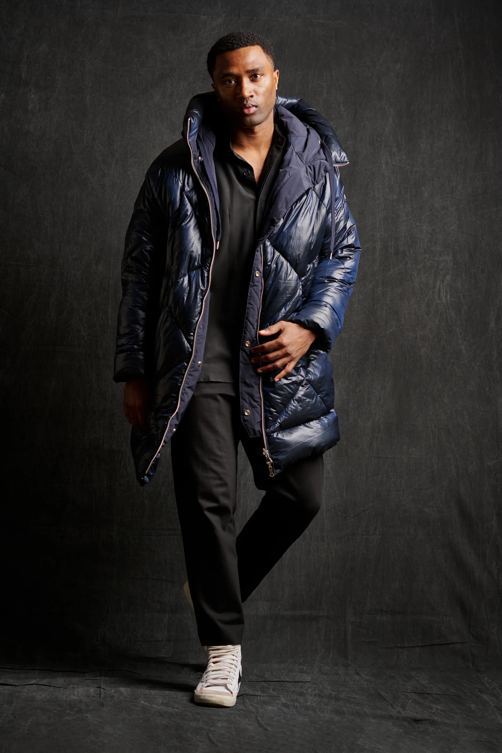 A model in a long dark blue padded coat and black trousers, standing with one foot crossed forward.