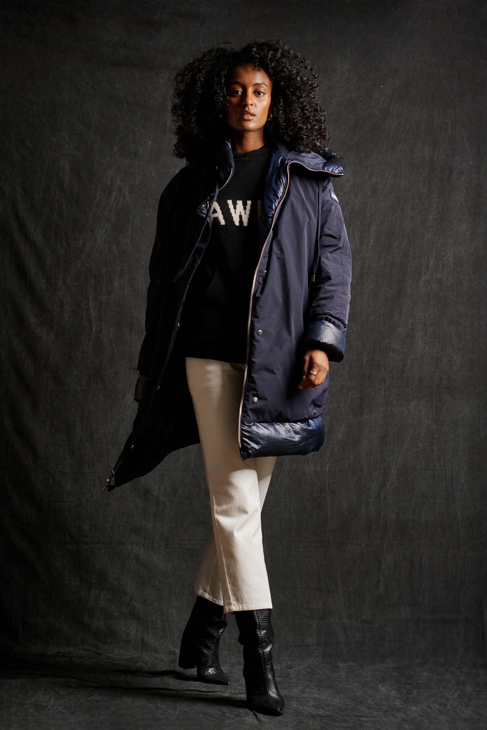 A model in a dark padded coat and cream trousers against a textured charcoal backdrop.