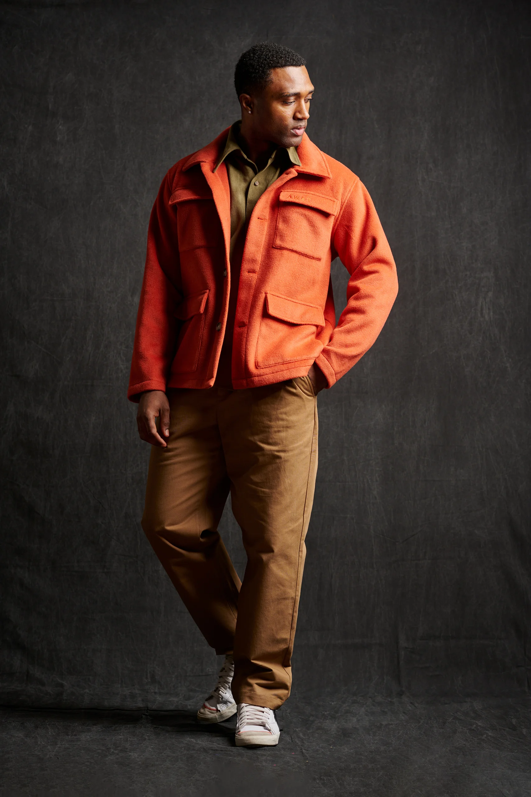 A model in an orange jacket and brown trousers, looking to the side.