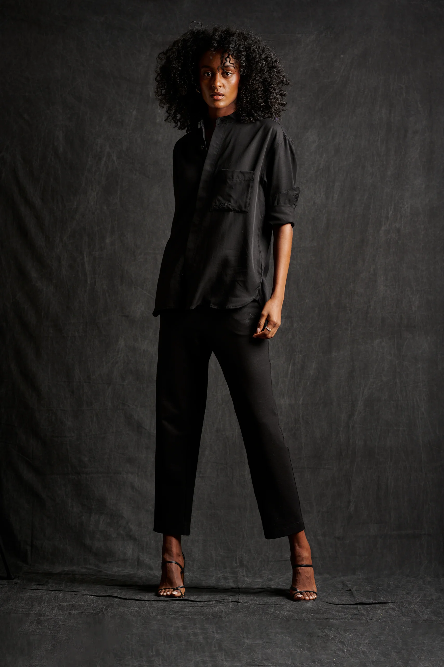 A model in a loose black shirt and cropped black trousers standing against charcoal.
