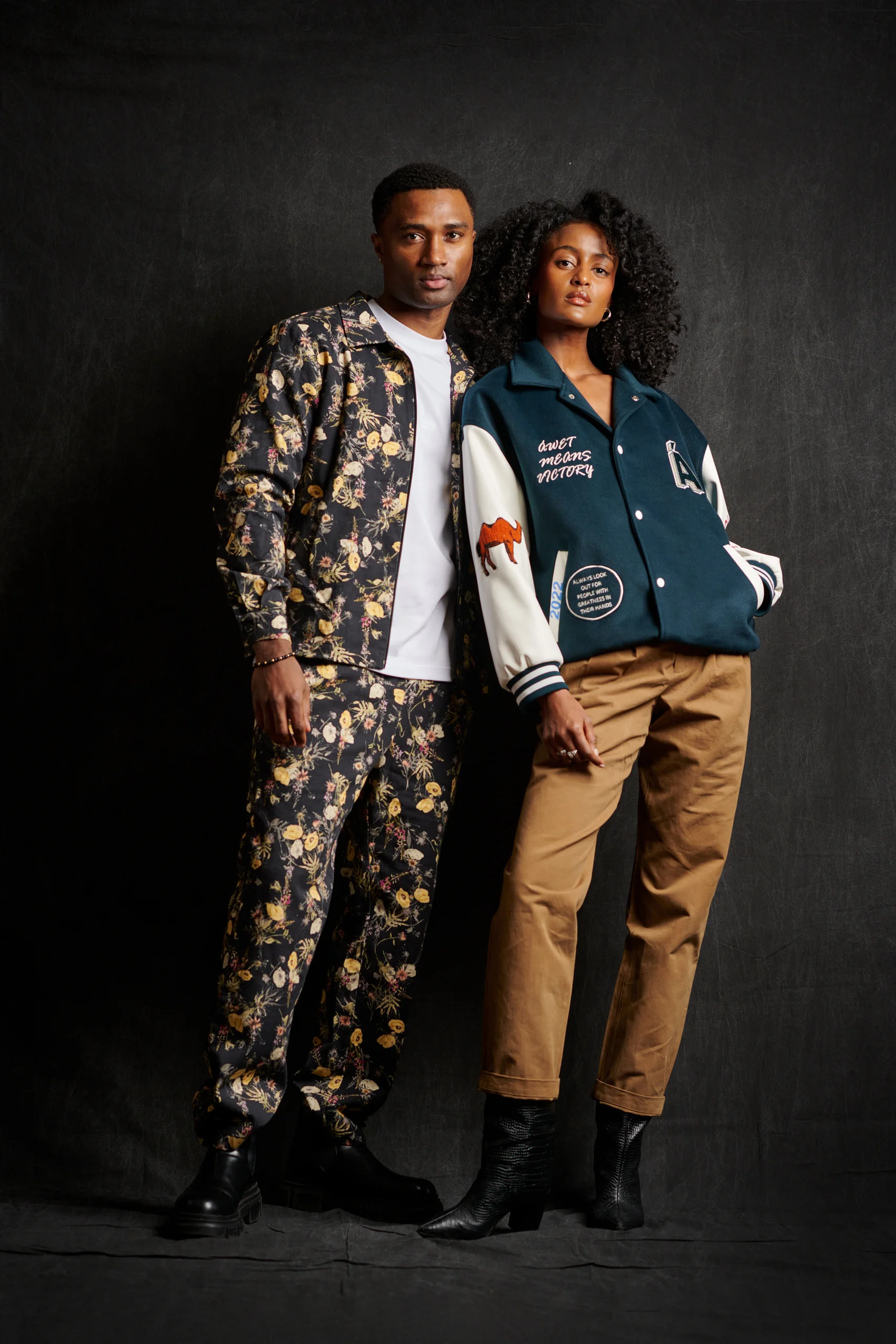 Two models standing close together in a patterned suit and a blue varsity jacket.
