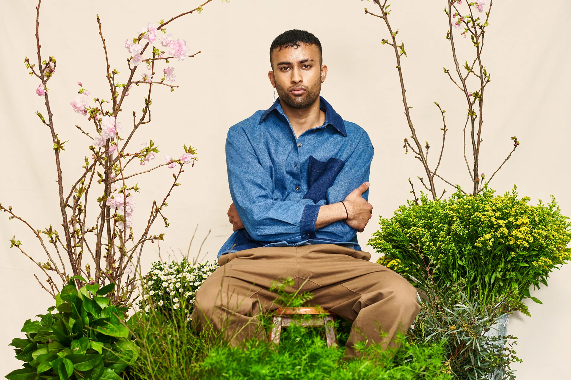 A seated model in a blue shirt and tan trousers among plants.
