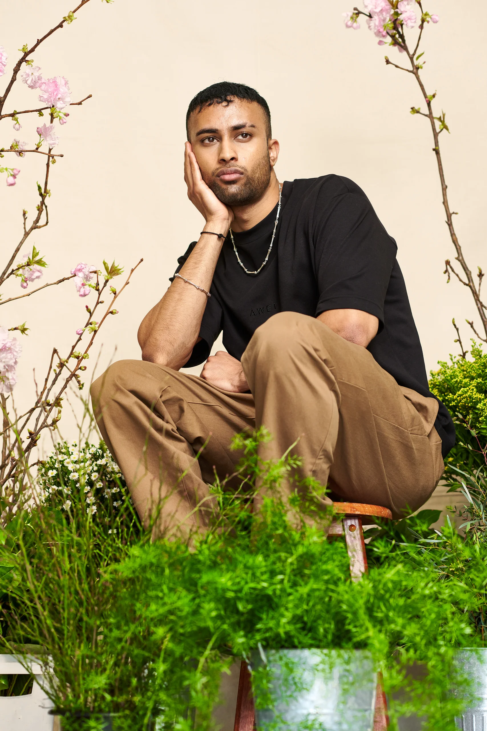 A seated model in a black shirt and tan trousers among greenery.