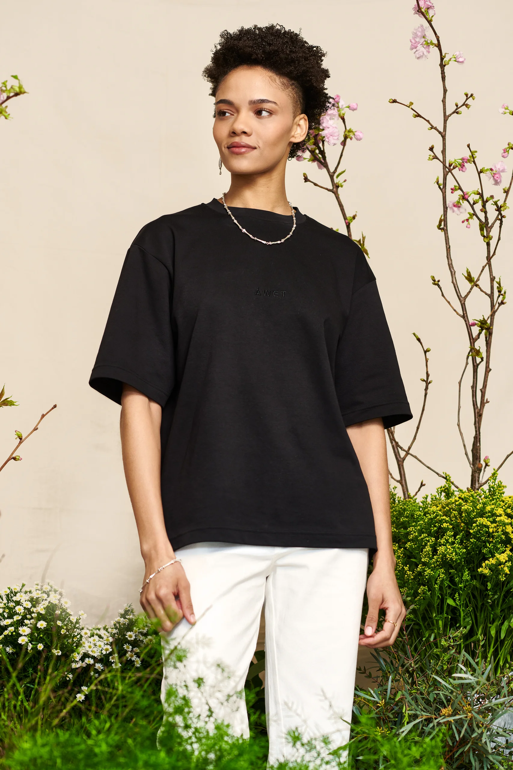 A model in a black shirt and white trousers framed by plants.