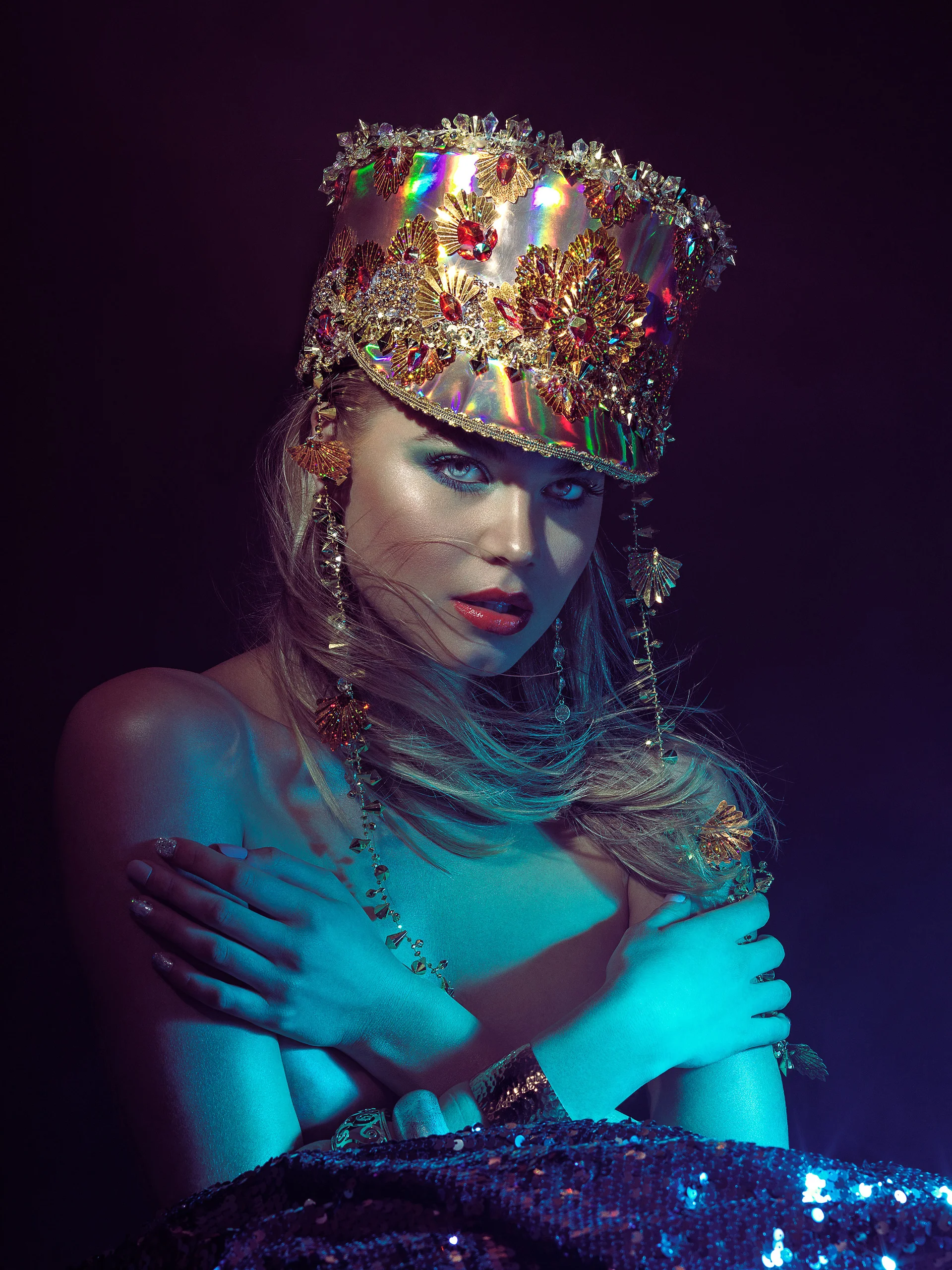 A model wearing a jeweled crown and layered jewelry under turquoise light.