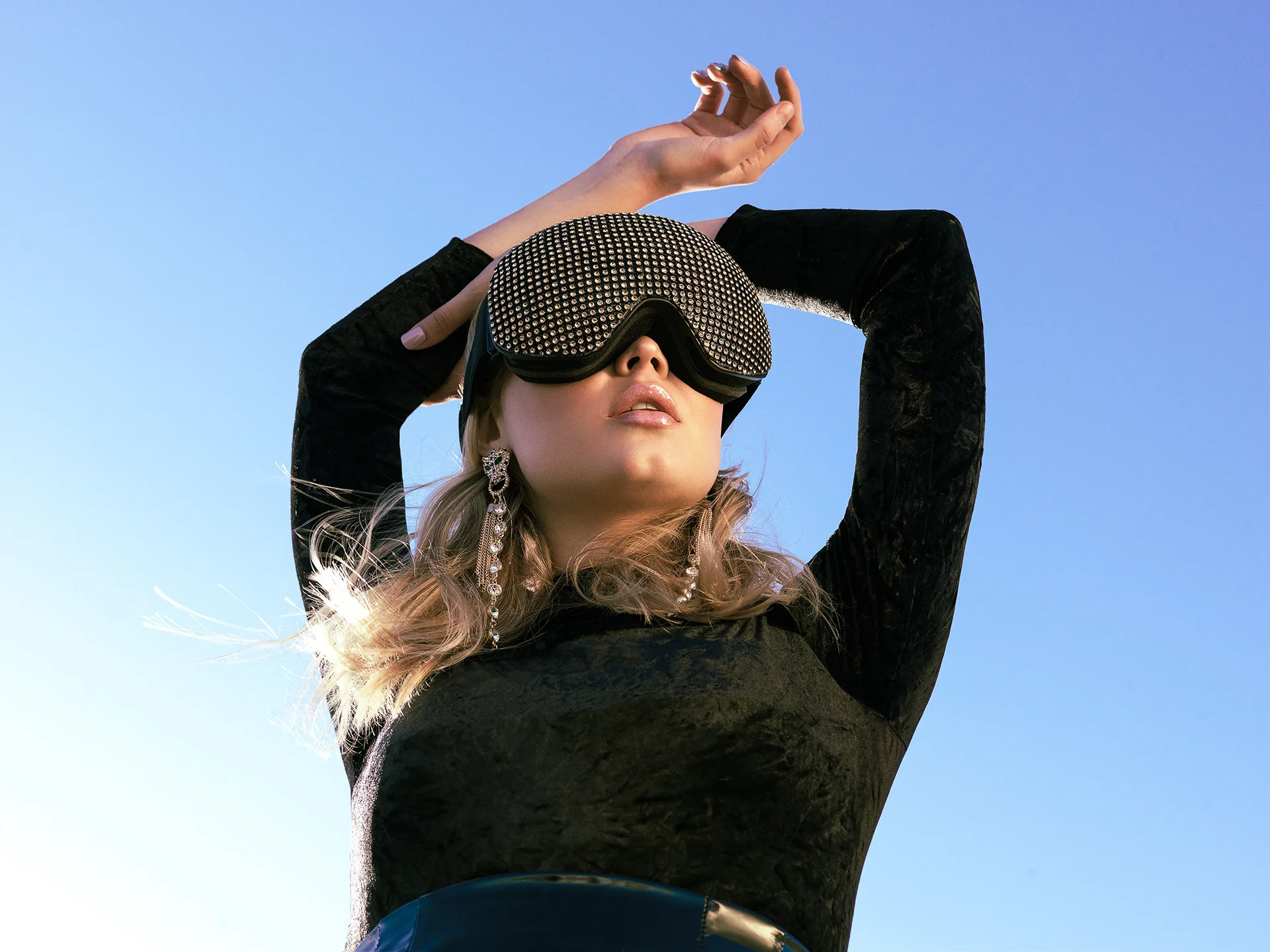A rooftop fashion portrait with oversized goggles and raised arms.