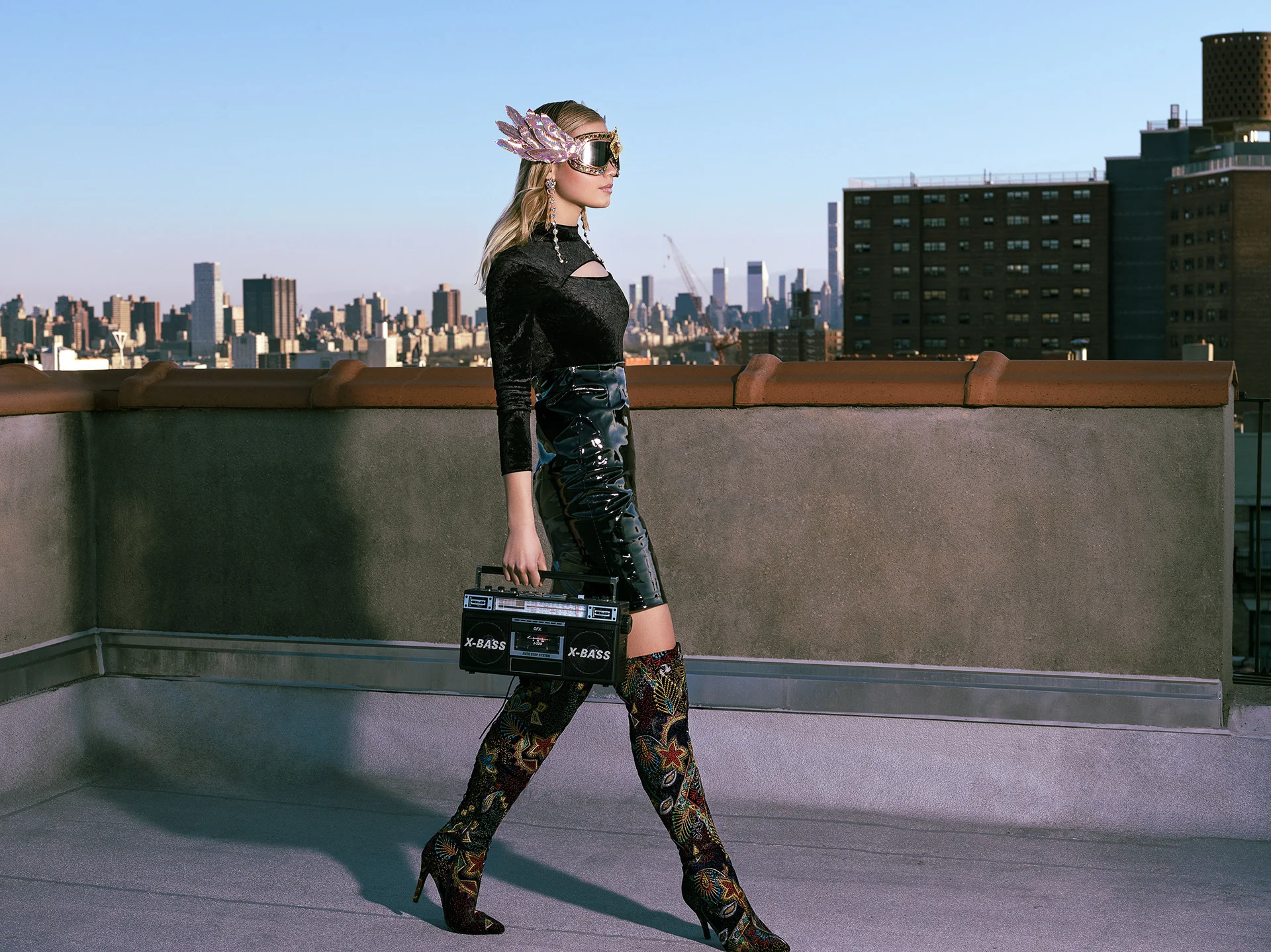 A model in black walking on a rooftop with a portable radio and skyline behind.