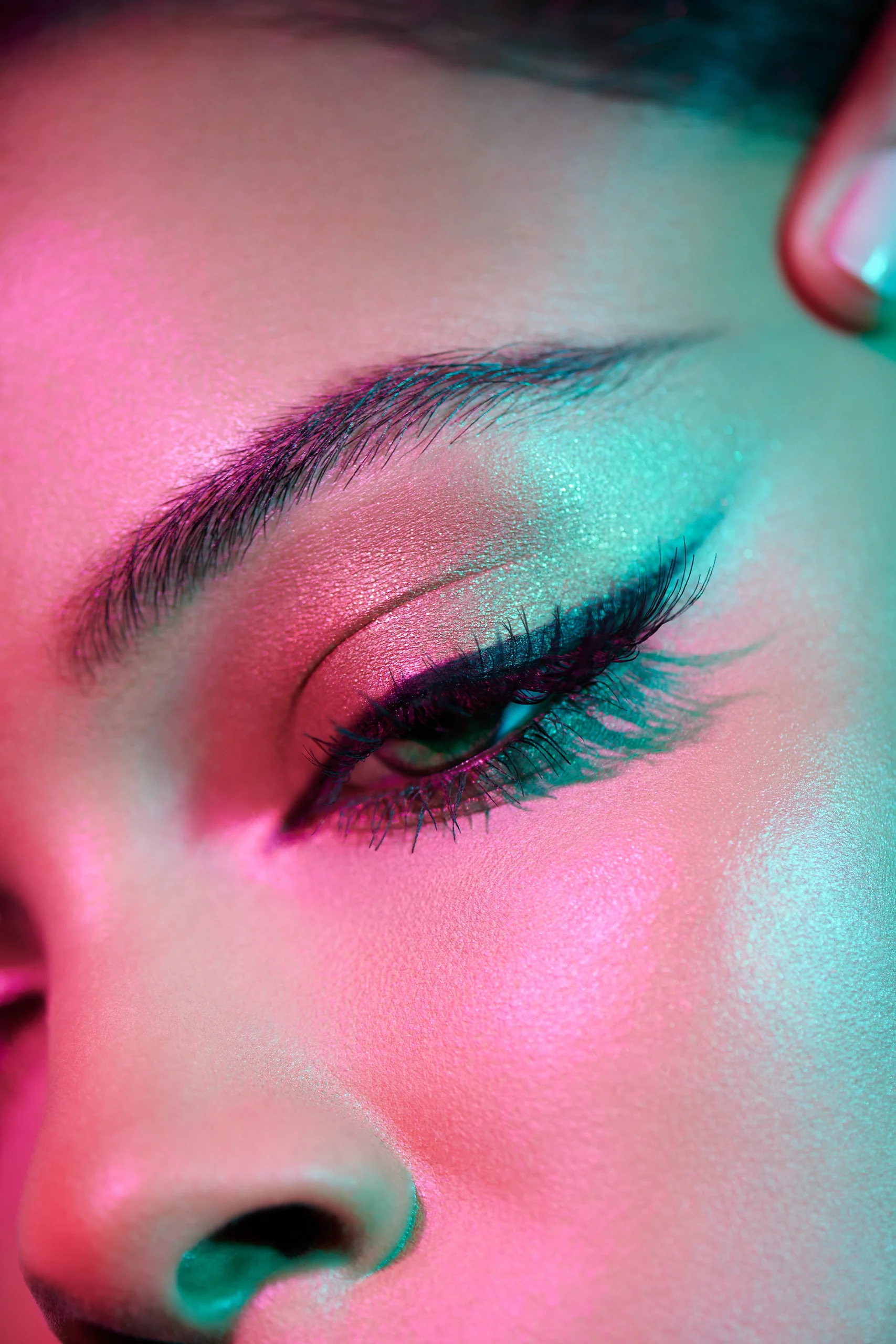 A close-up of winged eyeliner and shimmering eye makeup under pink and turquoise light.