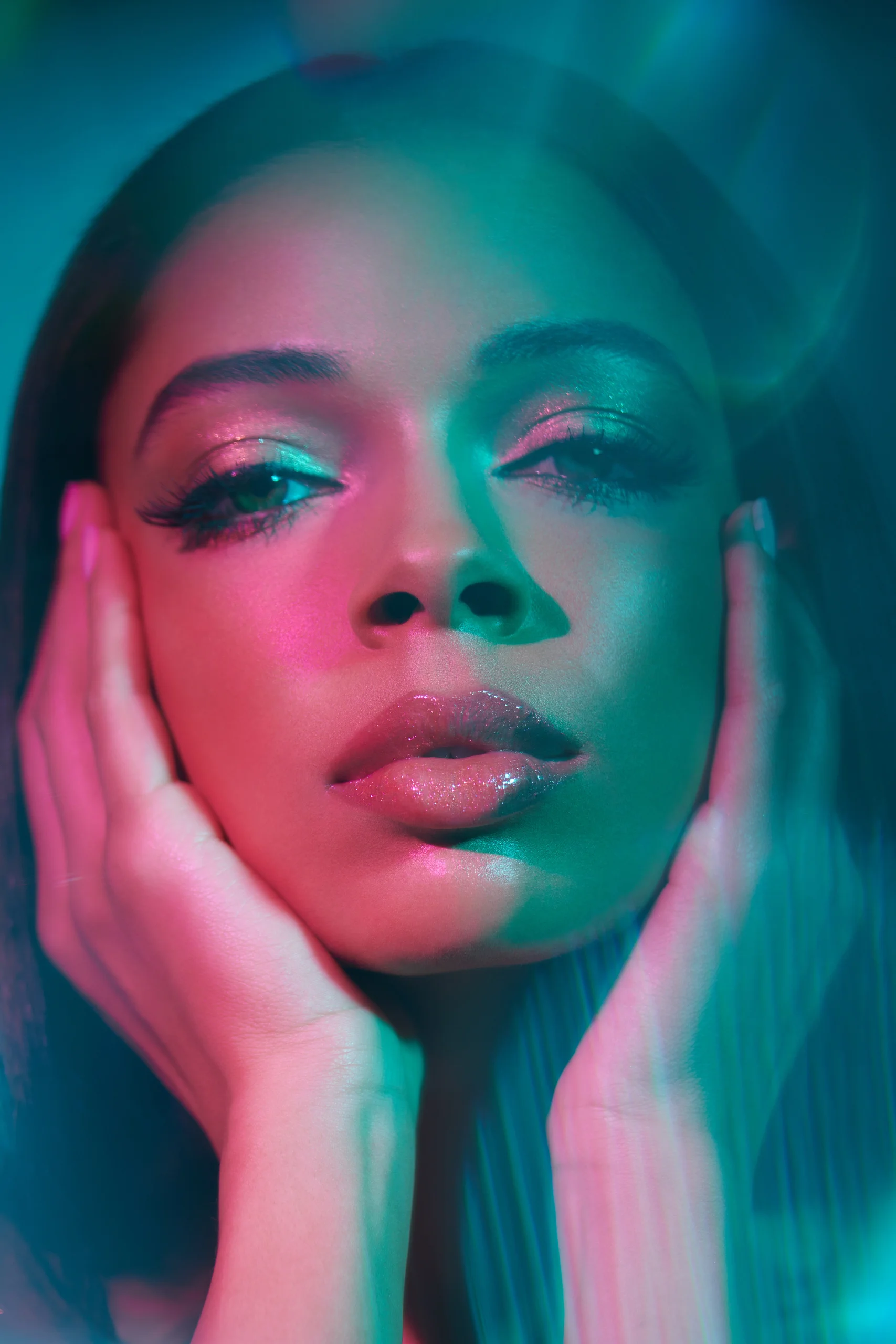 A model holds both hands against the face under turquoise and pink light.