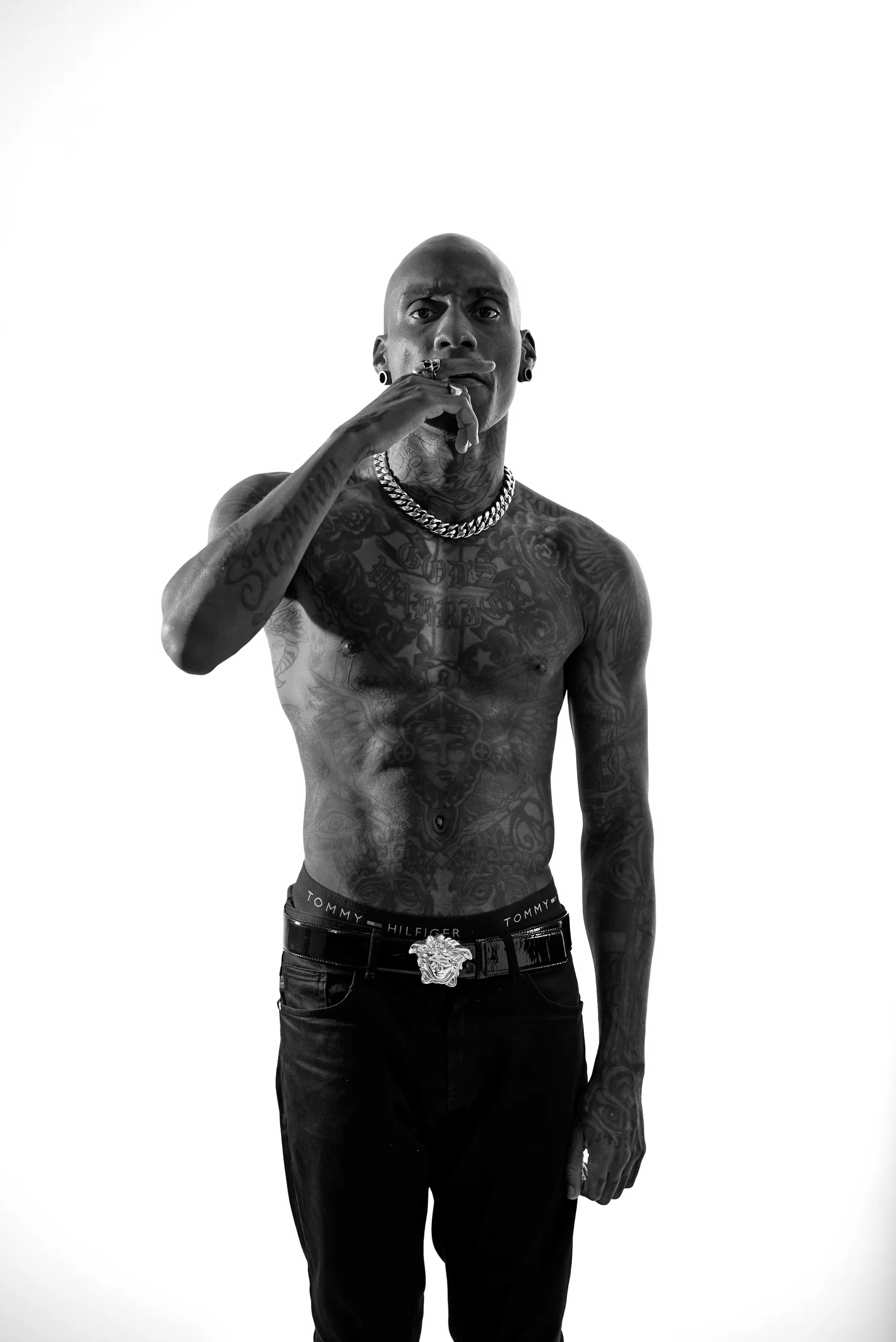 A black-and-white portrait of a bare-chested figure with one hand near the mouth.