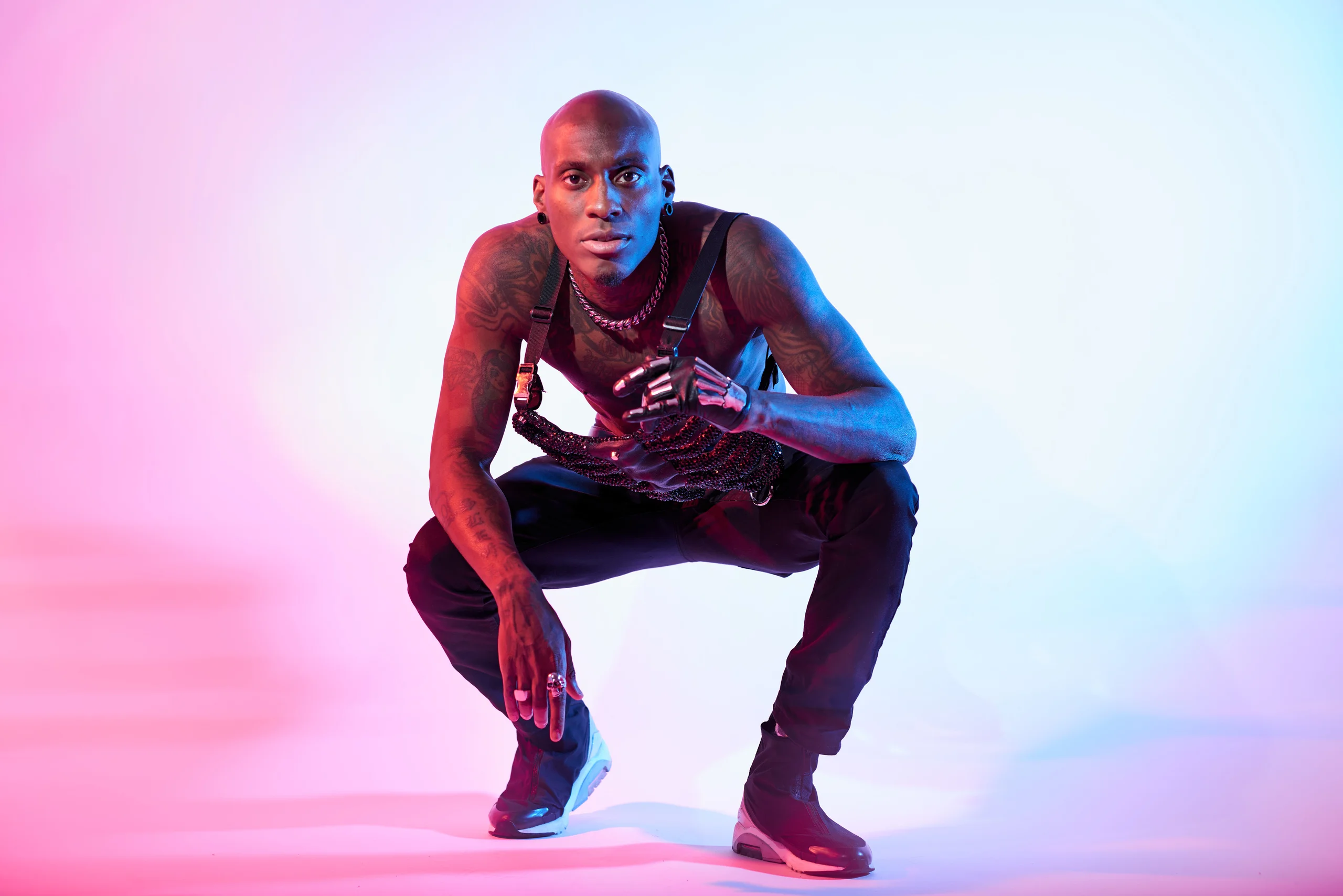 A crouching figure looking forward with one hand across the chest under pink and blue light.