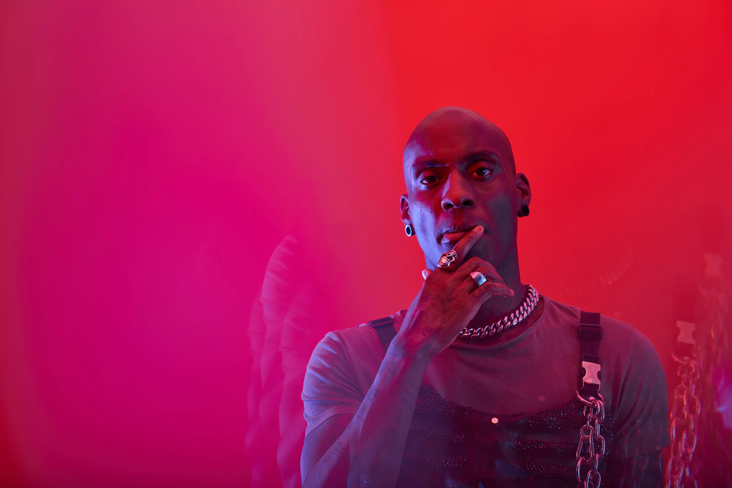 A close portrait with a chain near the mouth against a red and magenta background.