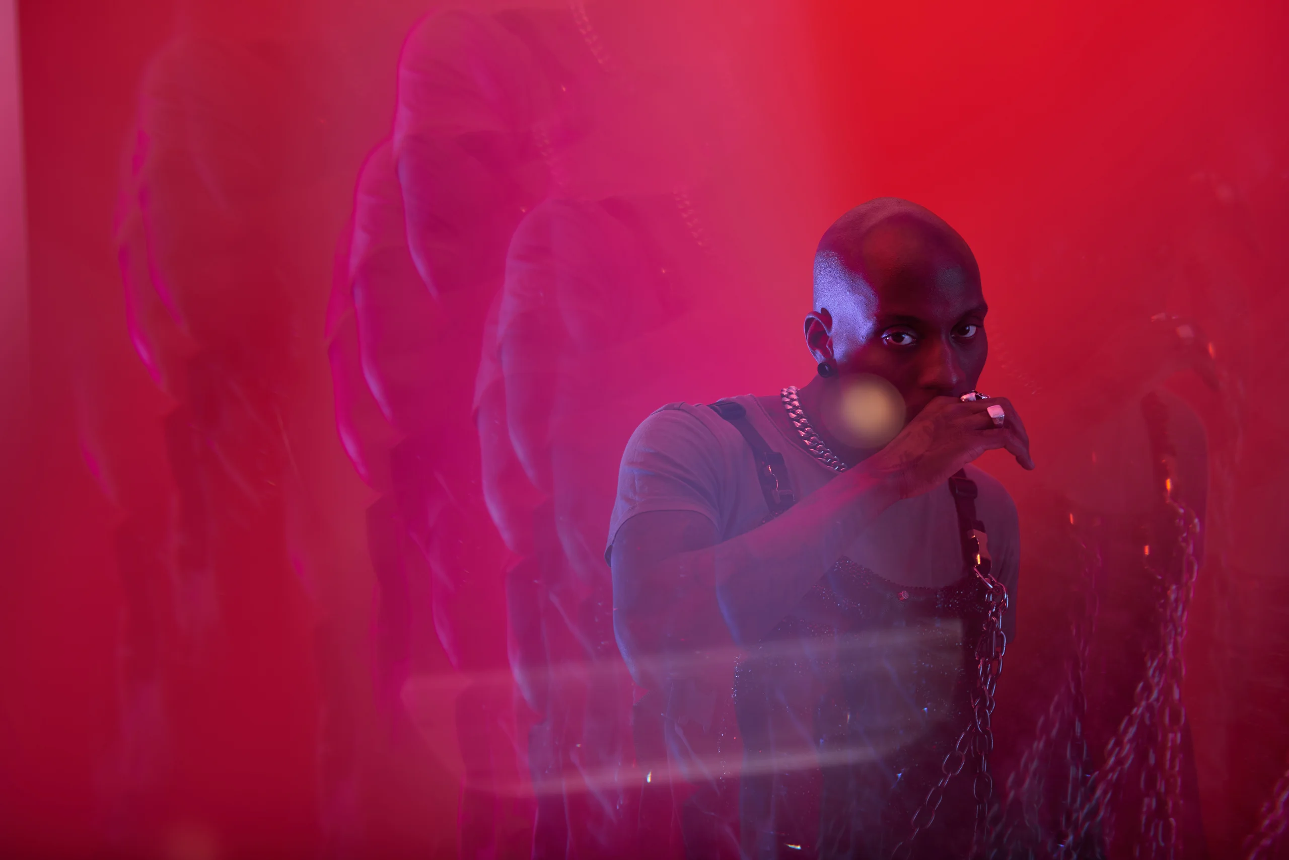 A figure behind layers of translucent red reflections, with a hand near the mouth.