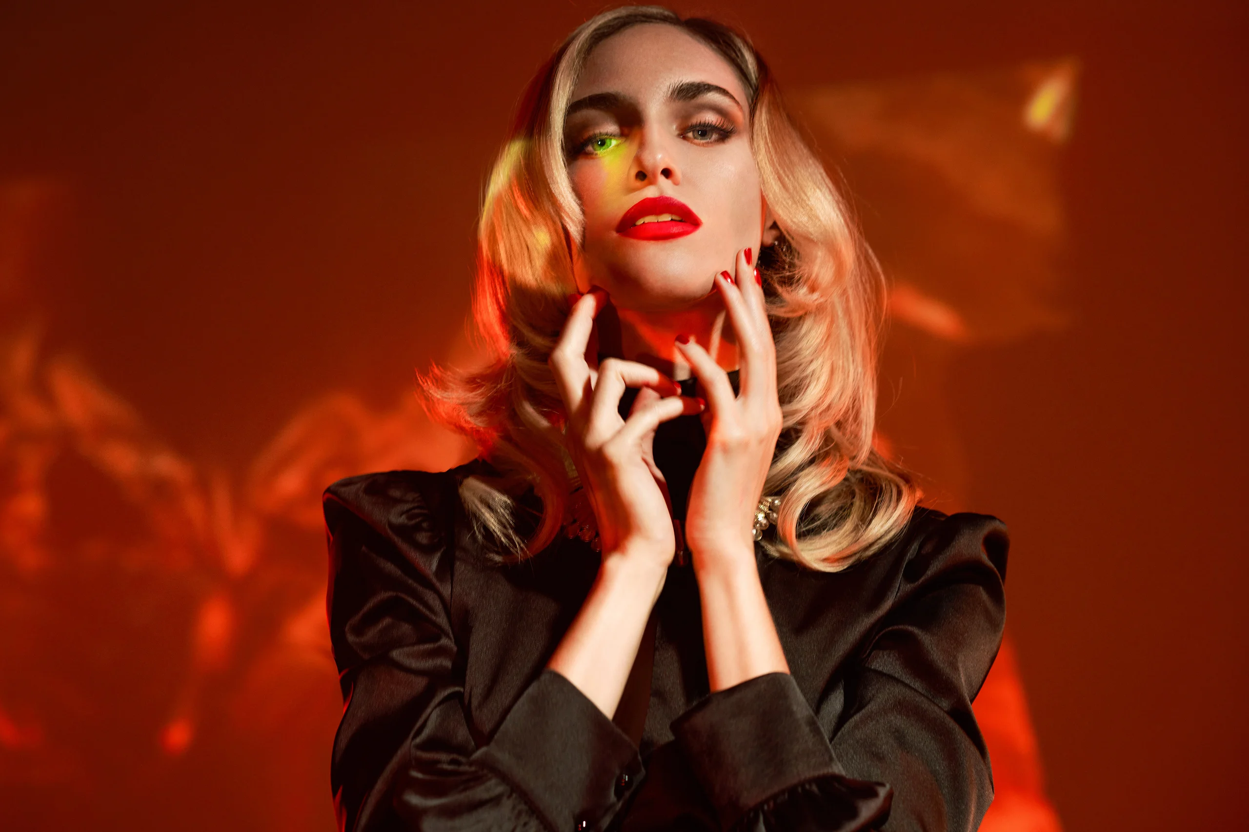 A model in black with red lips and hands at the neck against projected orange light.