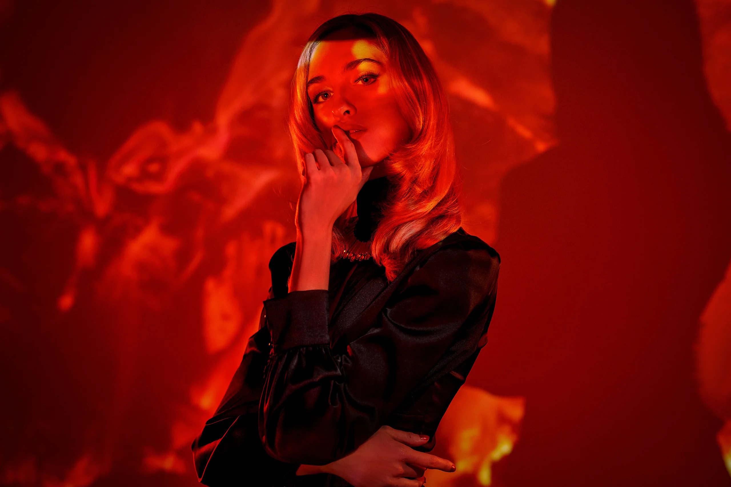 A seated model in a black tailored outfit under red and orange projected light.