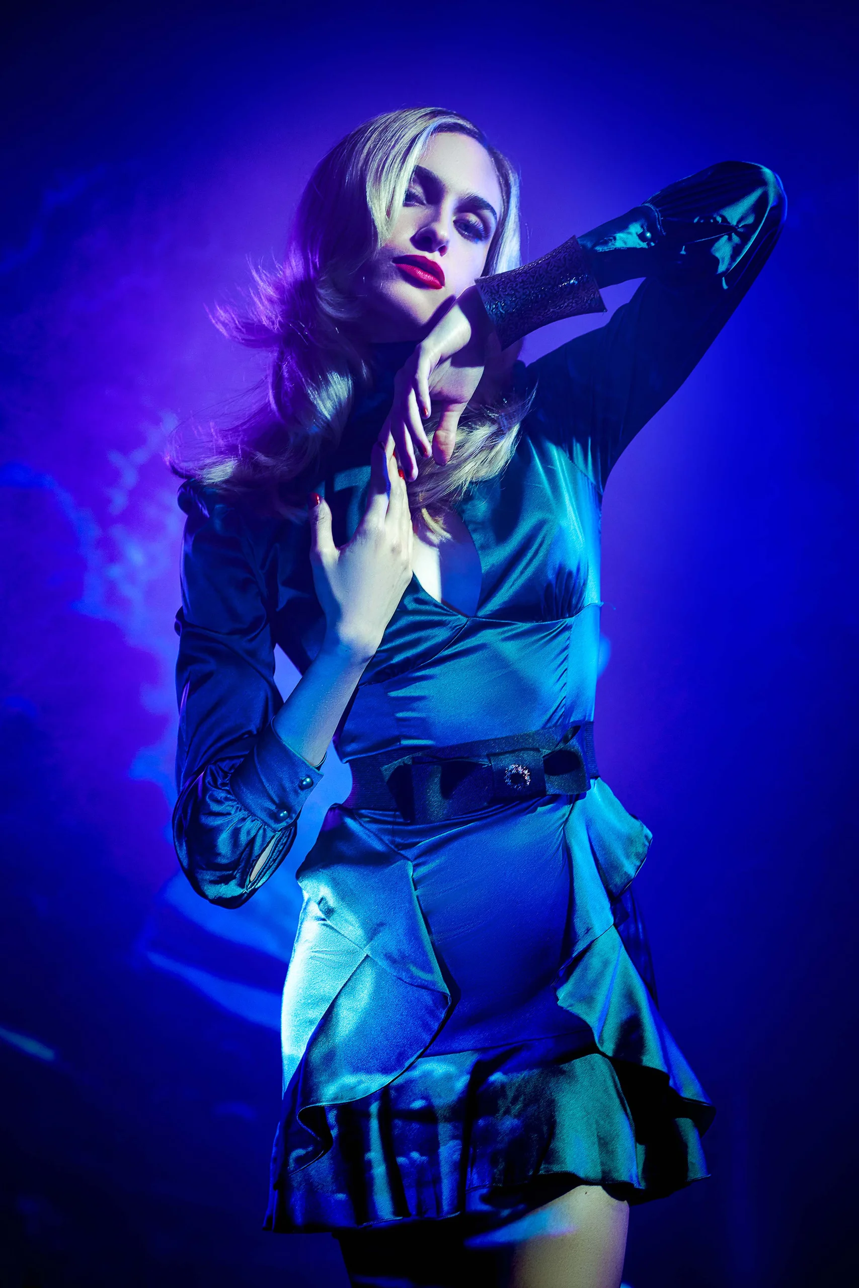 A model in a blue dress raising an arm beneath violet and blue light.