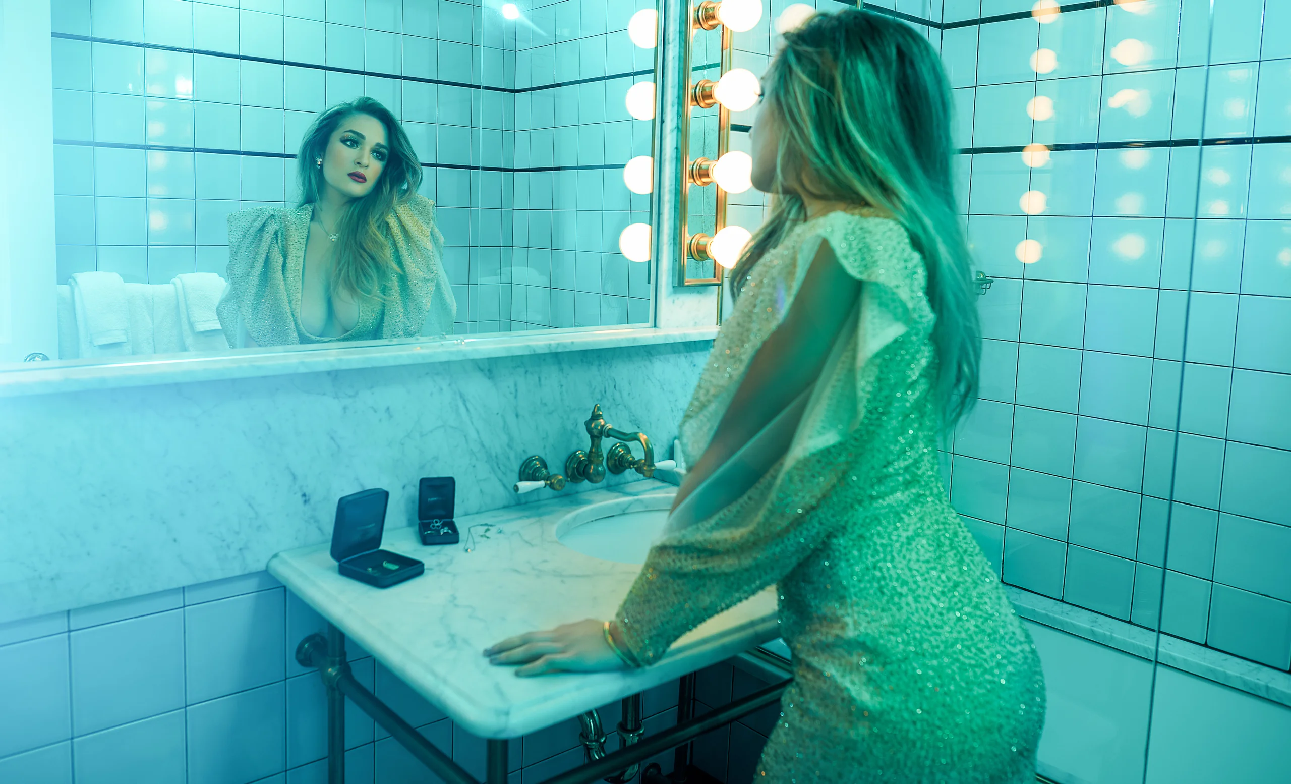 A model in a metallic dress leaning over a tiled sink and looking into the mirror.