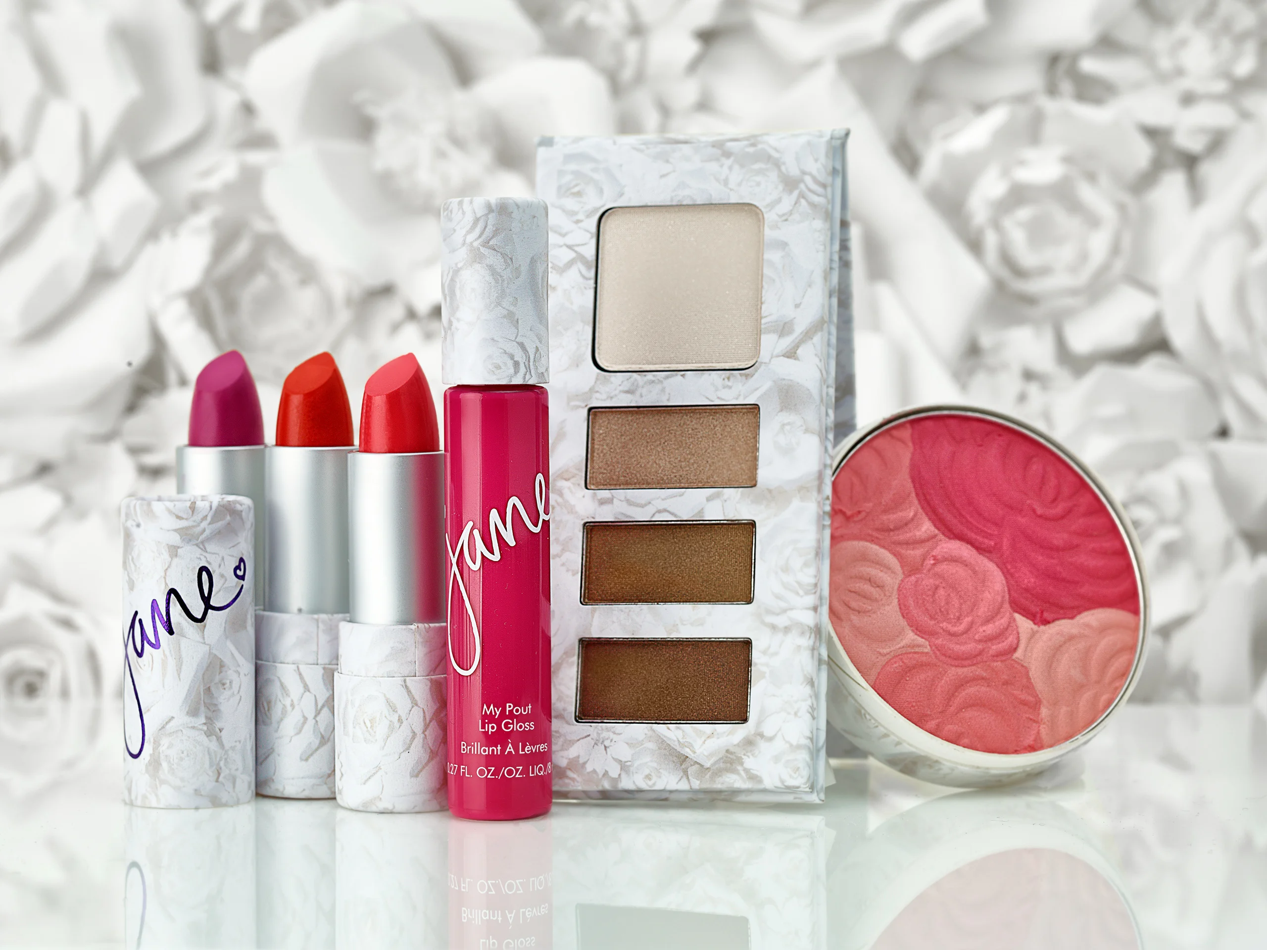 Jane lipsticks, lip gloss, blush, and an eye palette arranged against a white floral backdrop.