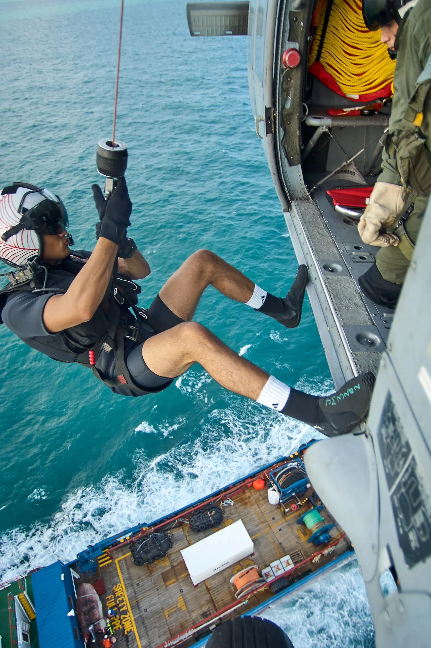A helmeted person suspended beside a helicopter above water and a boat.
