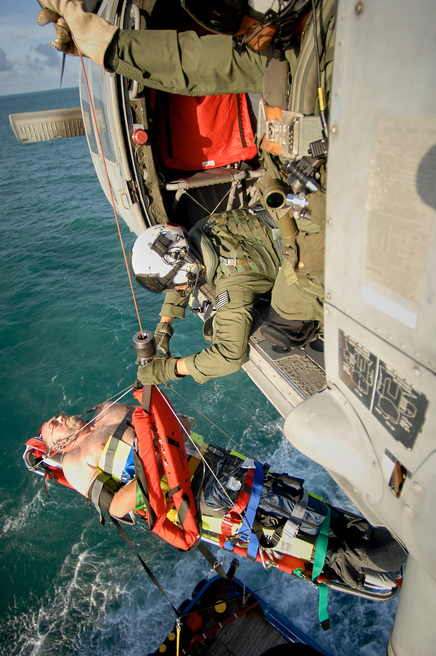 A person secured on an orange stretcher being lifted beside a helicopter over water.