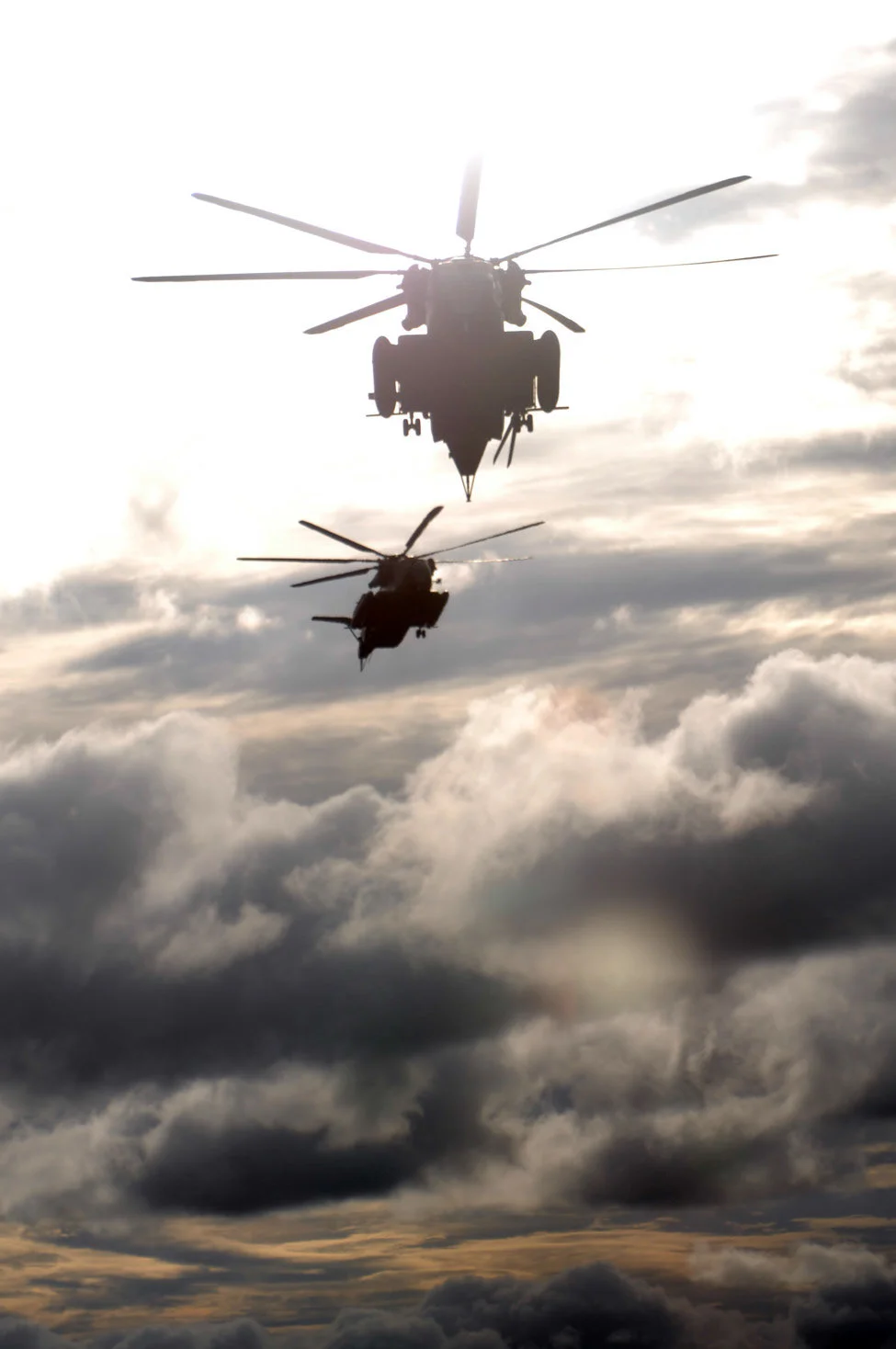 Two helicopter silhouettes above clouds in bright backlight.