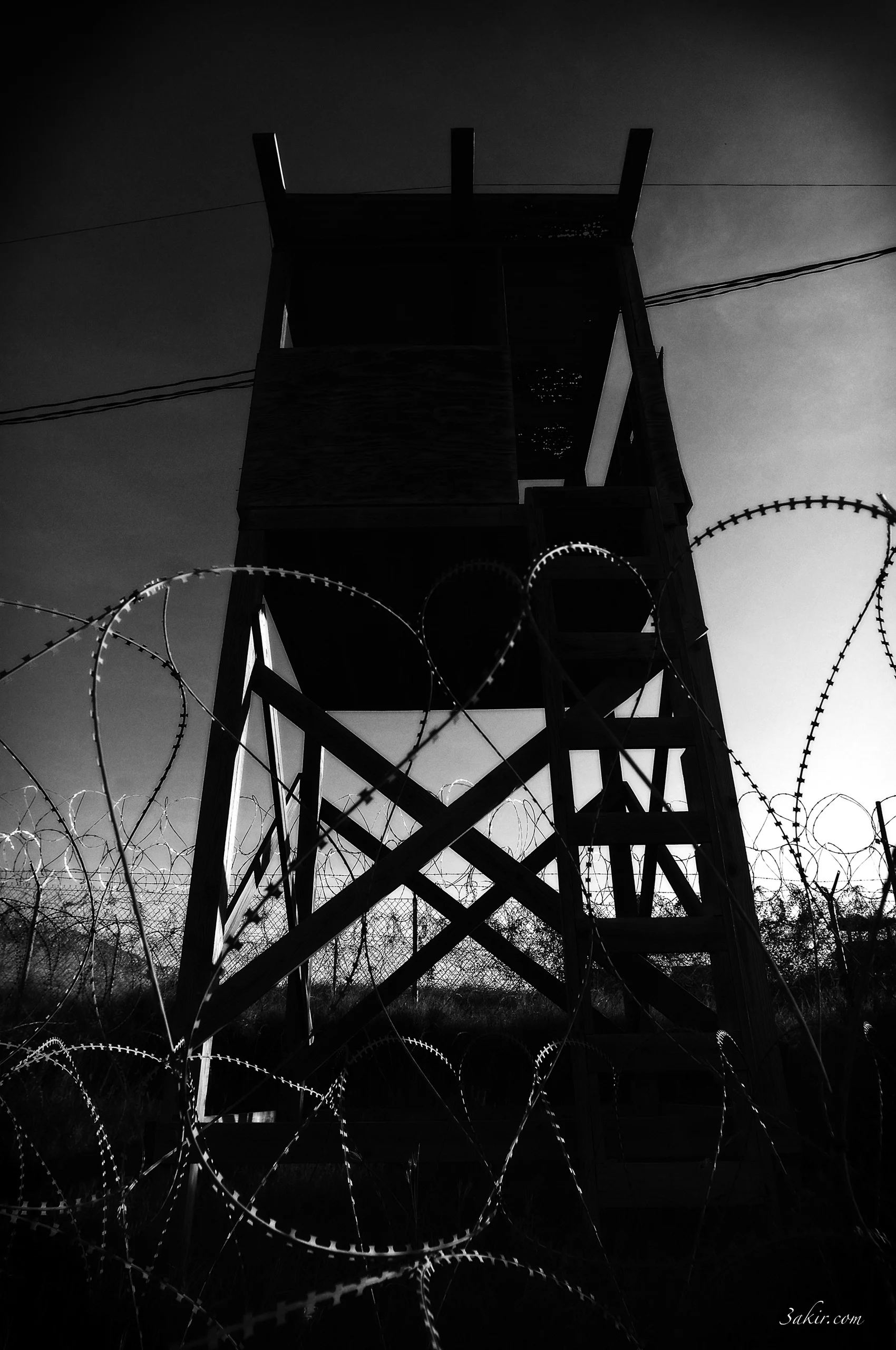 A black-and-white view of a watchtower framed by coils of barbed wire.