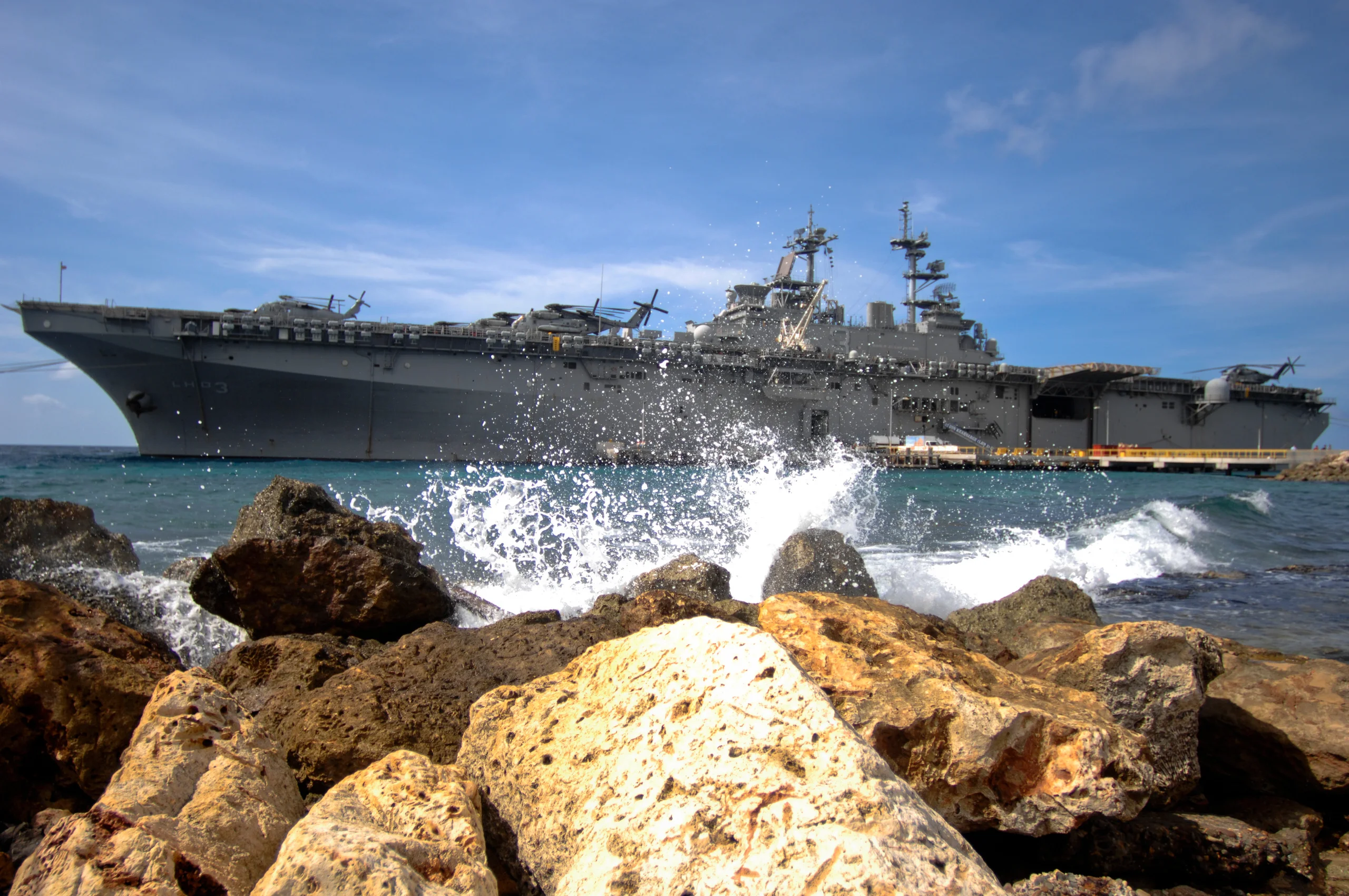 A large naval ship offshore beyond rocks and breaking waves.