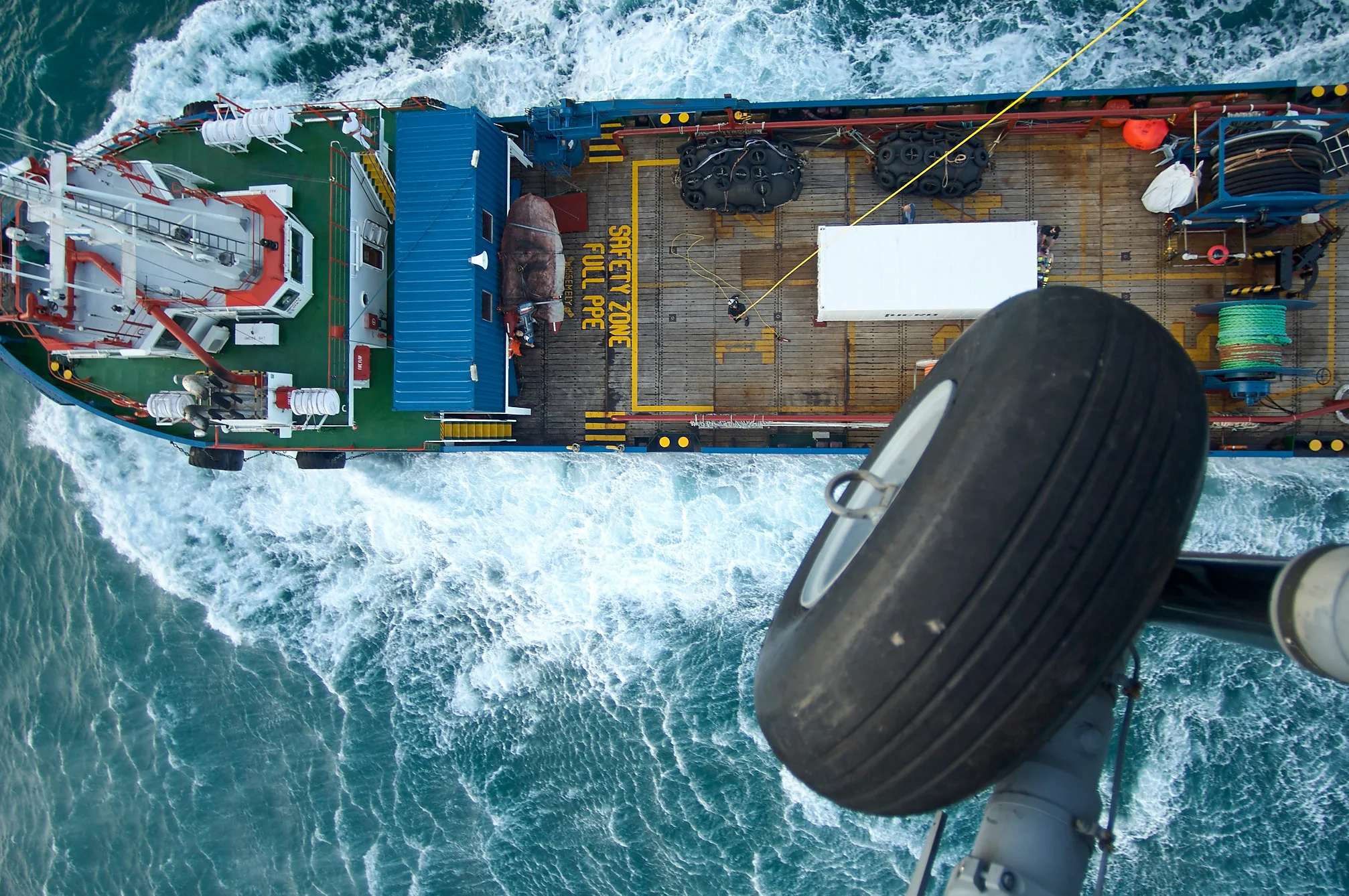 An overhead view of a boat beneath an aircraft wheel above churning water.