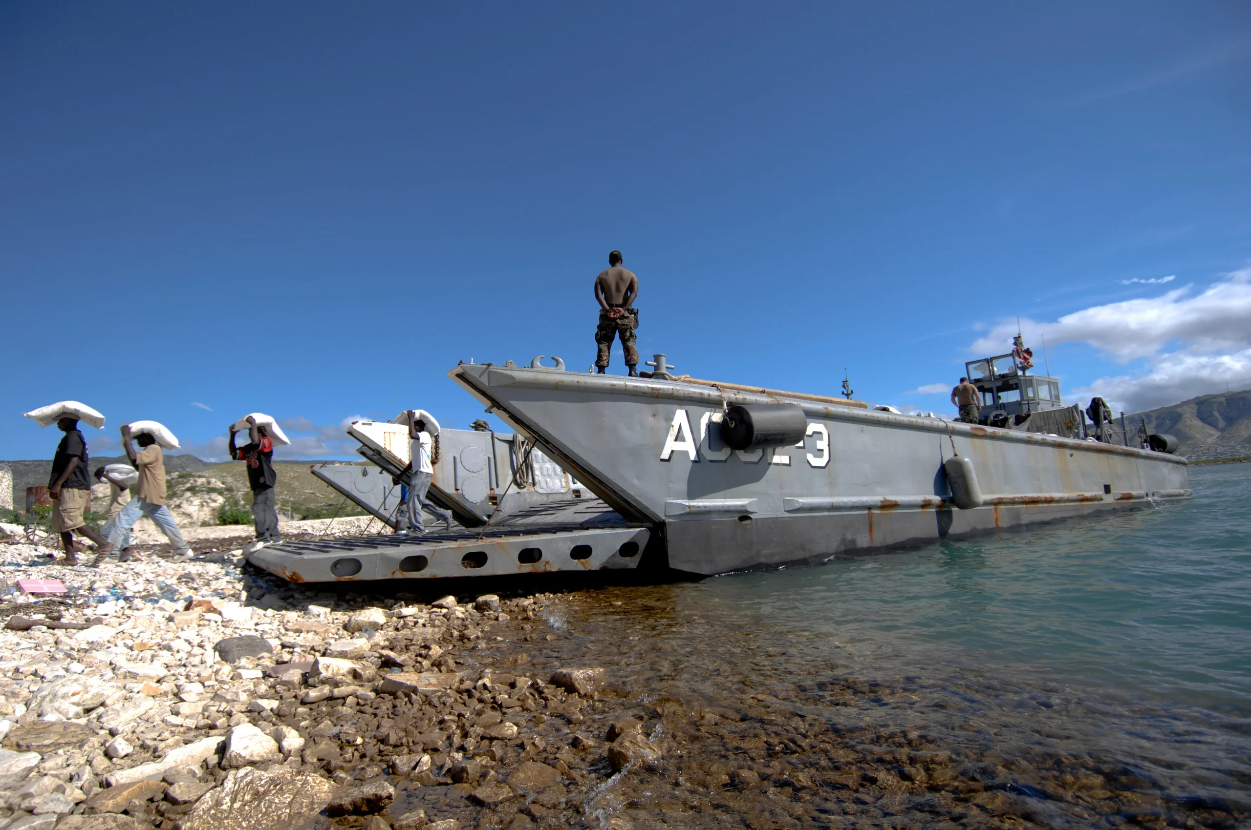 People unloading supplies from a landing craft onto a rocky shore.