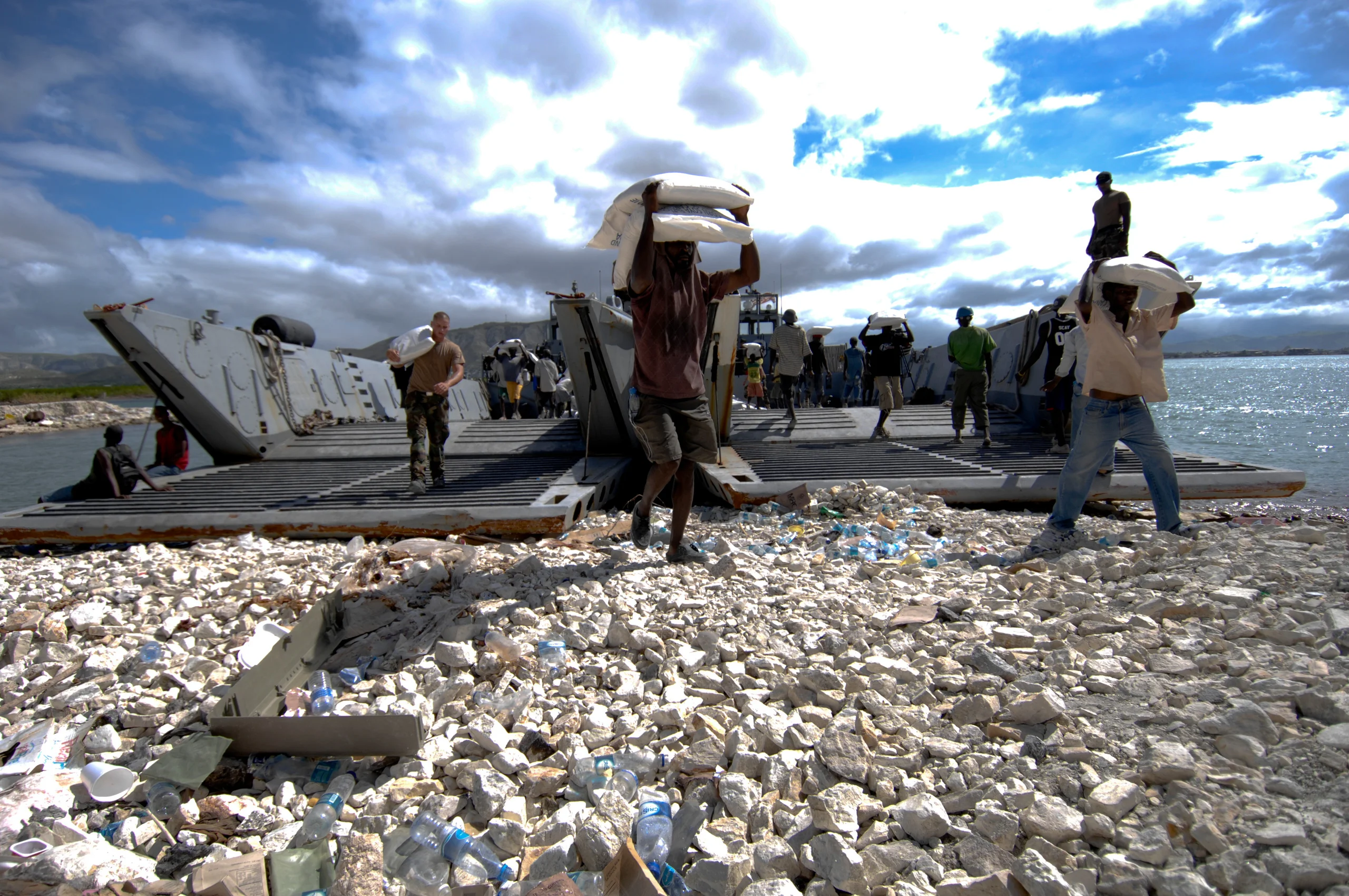 People carrying boxes away from a landing craft on a rocky beach.