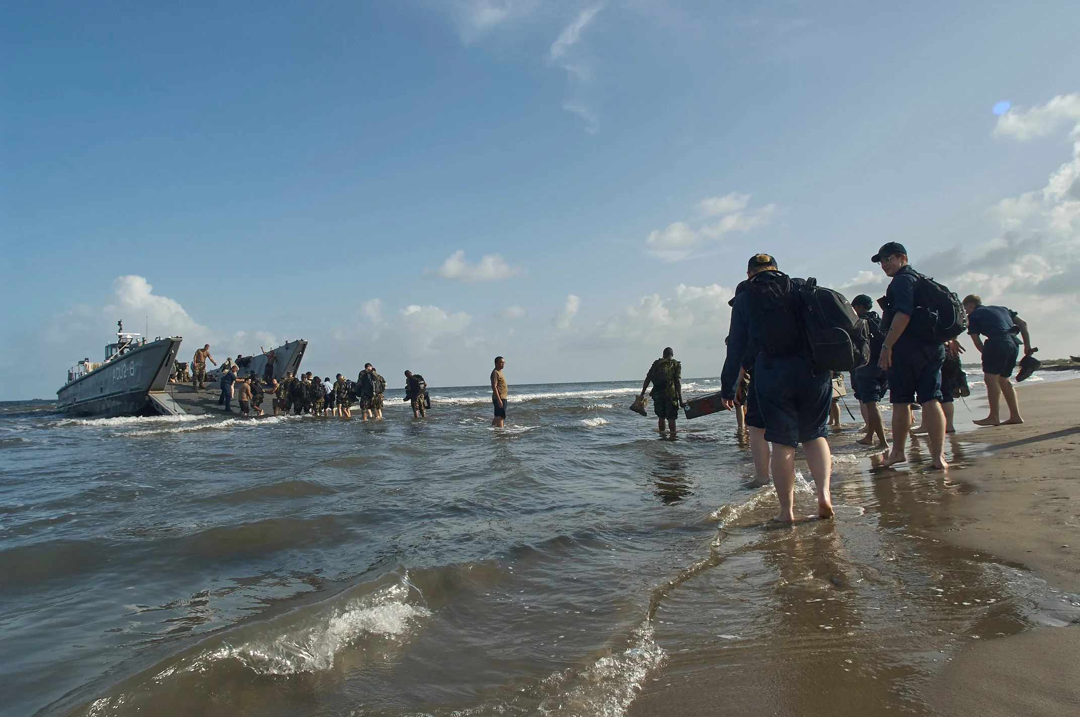 People carrying bags through shallow water toward a beach, with a landing craft behind them.