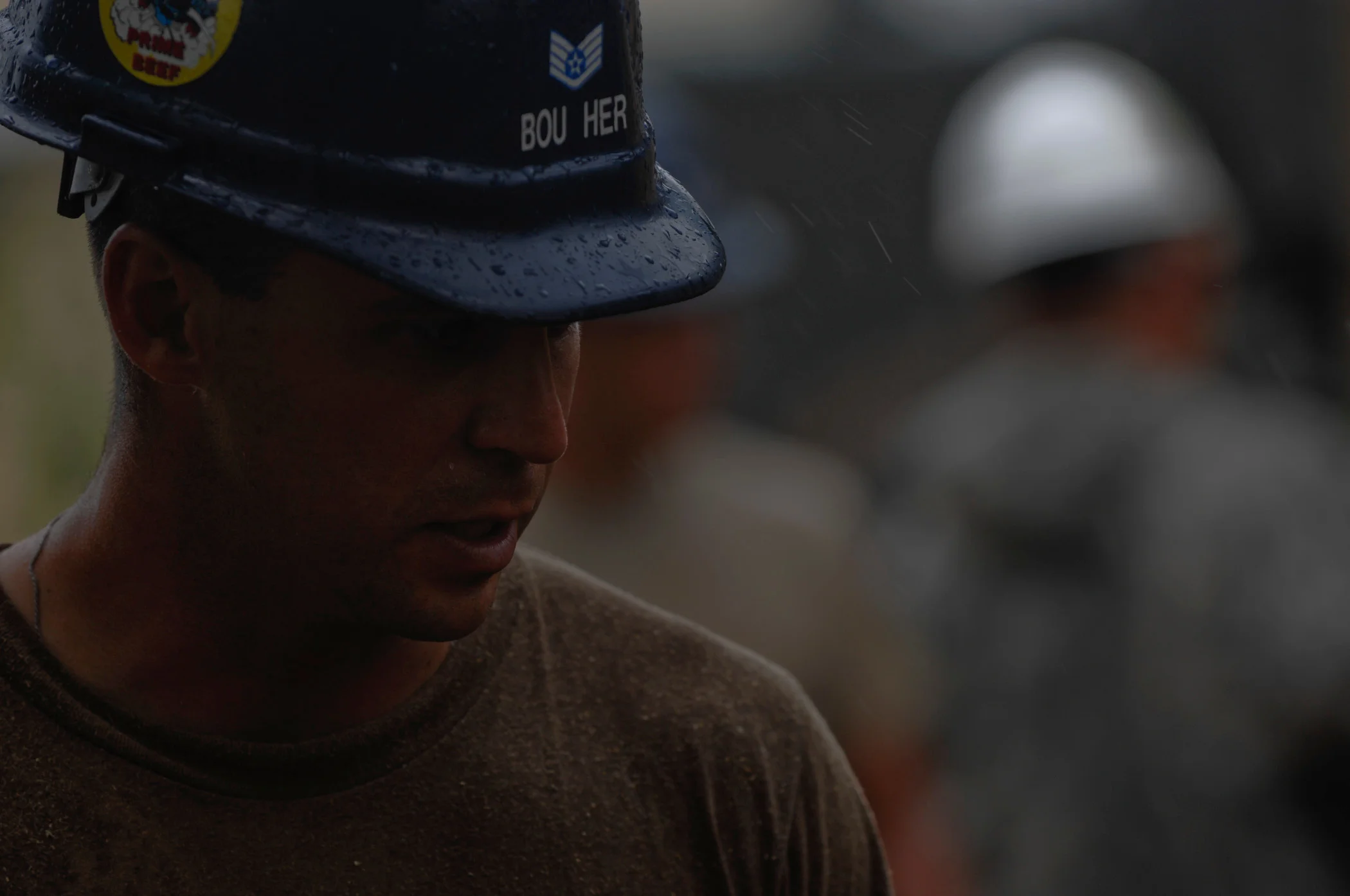 A close portrait of a person wearing a hard hat, with another figure blurred behind.