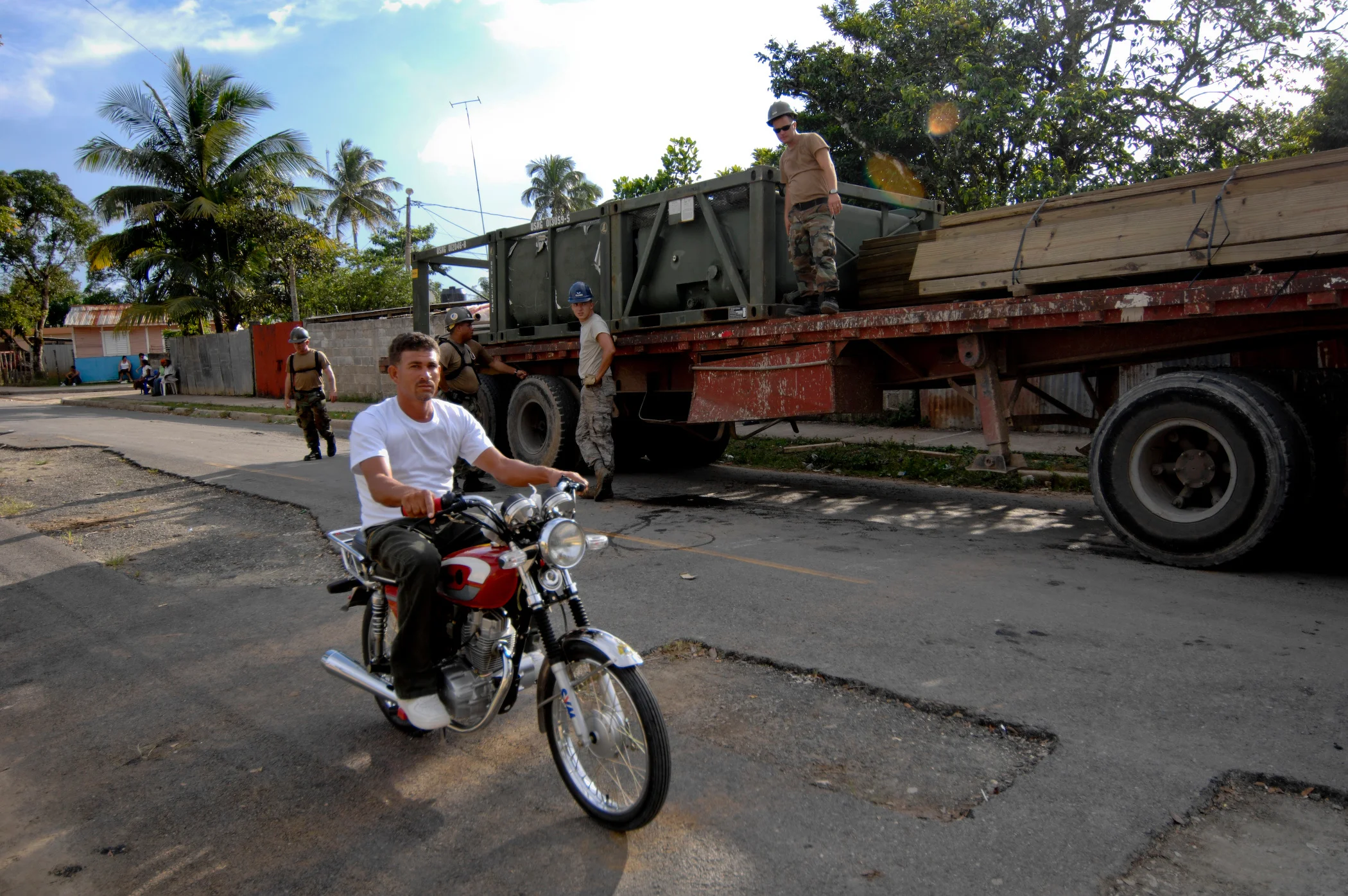 A motorcyclist passing a parked flatbed truck on a palm-lined street.