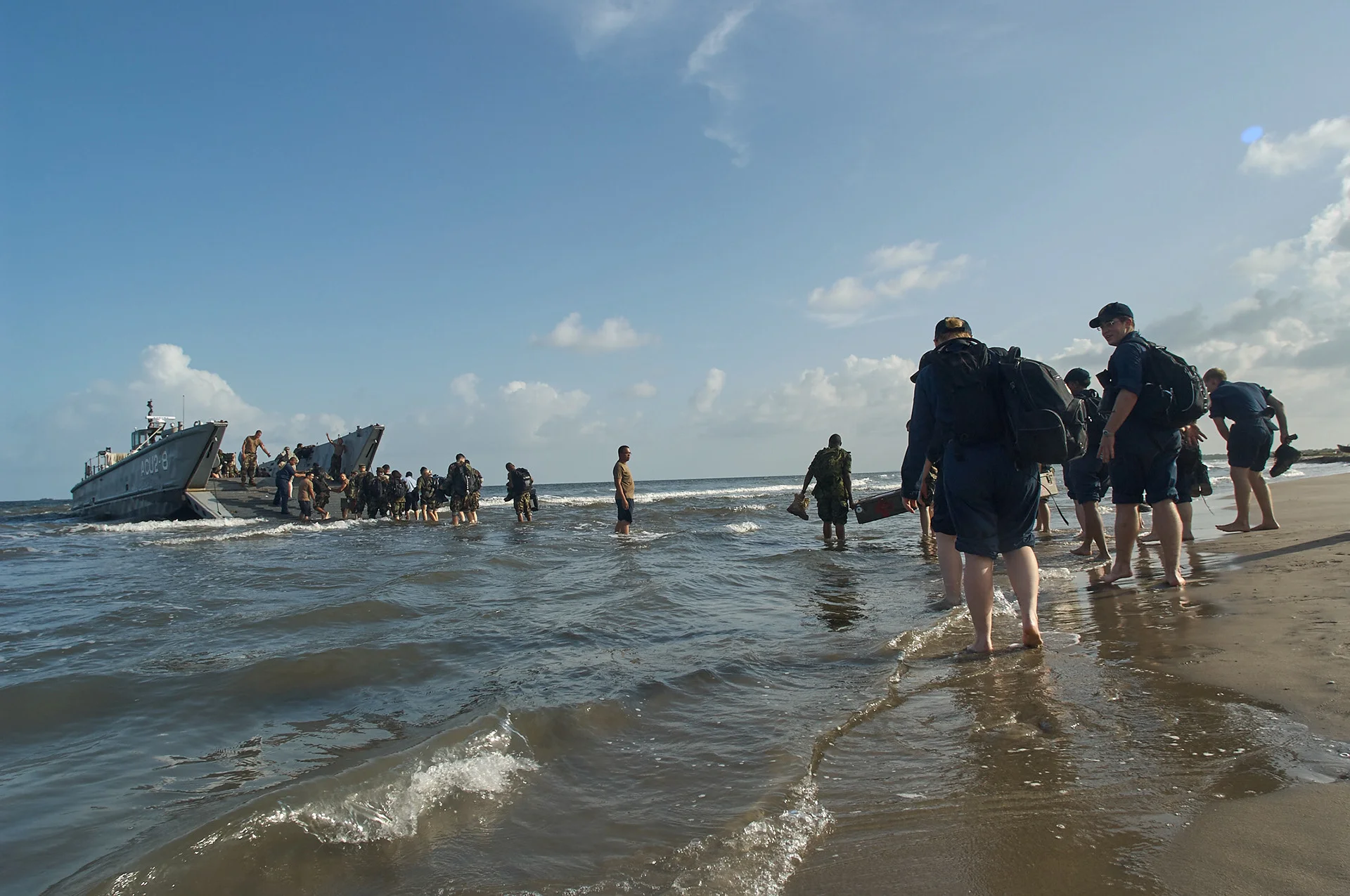 People carrying bags through shallow water near a landing craft and sandy shore.