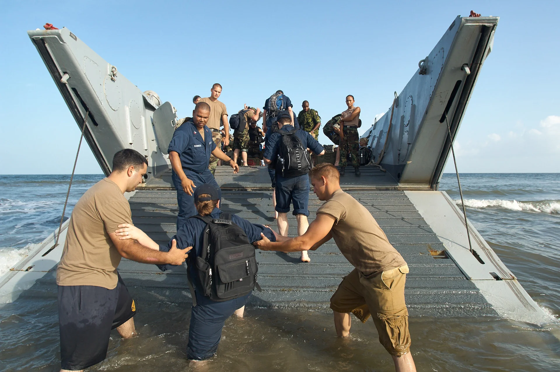 People helping someone down a landing craft ramp into shallow water.