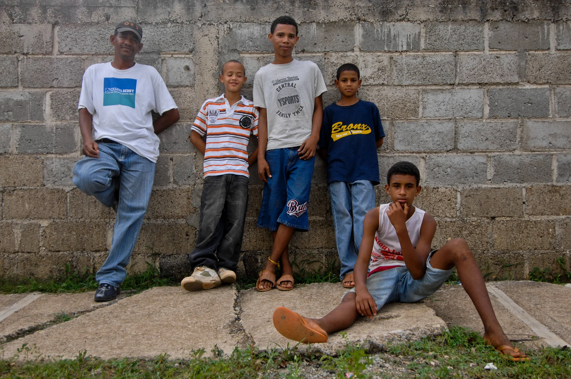 Five people posing beside a concrete block wall, with one seated on the ground.